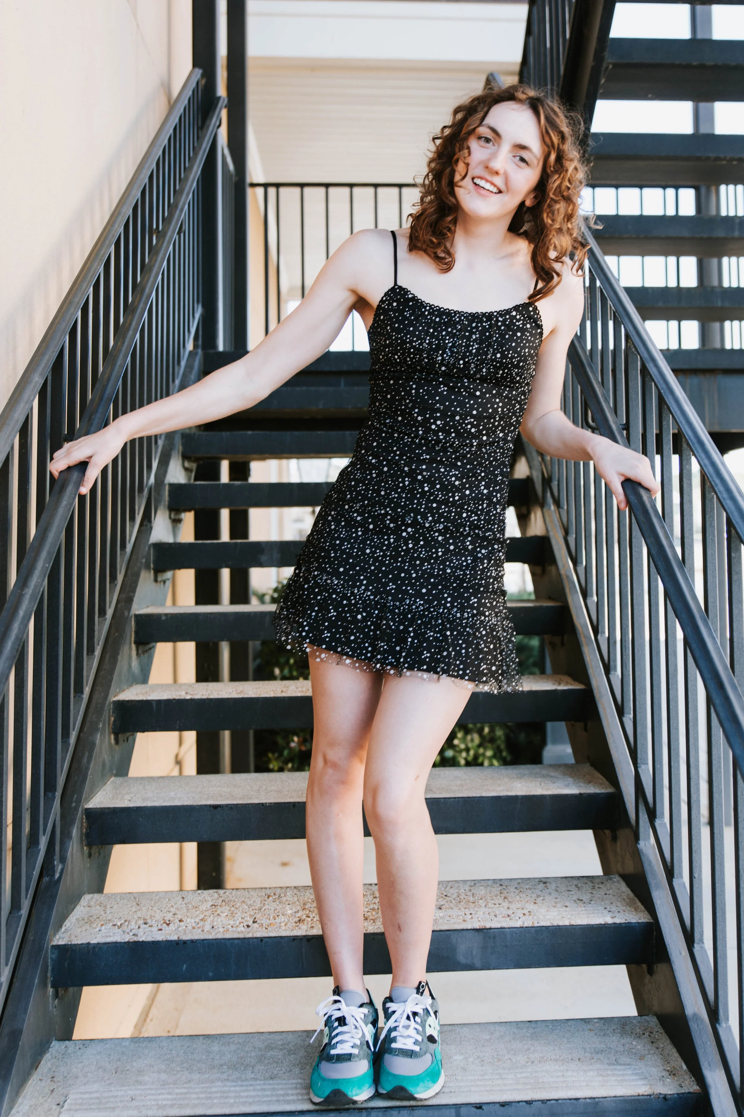 A young woman with curly hair smiling, wearing a black dress with white polka dots and sneakers, standing on an outdoor staircase.  Senior Photographer Madison, MS 