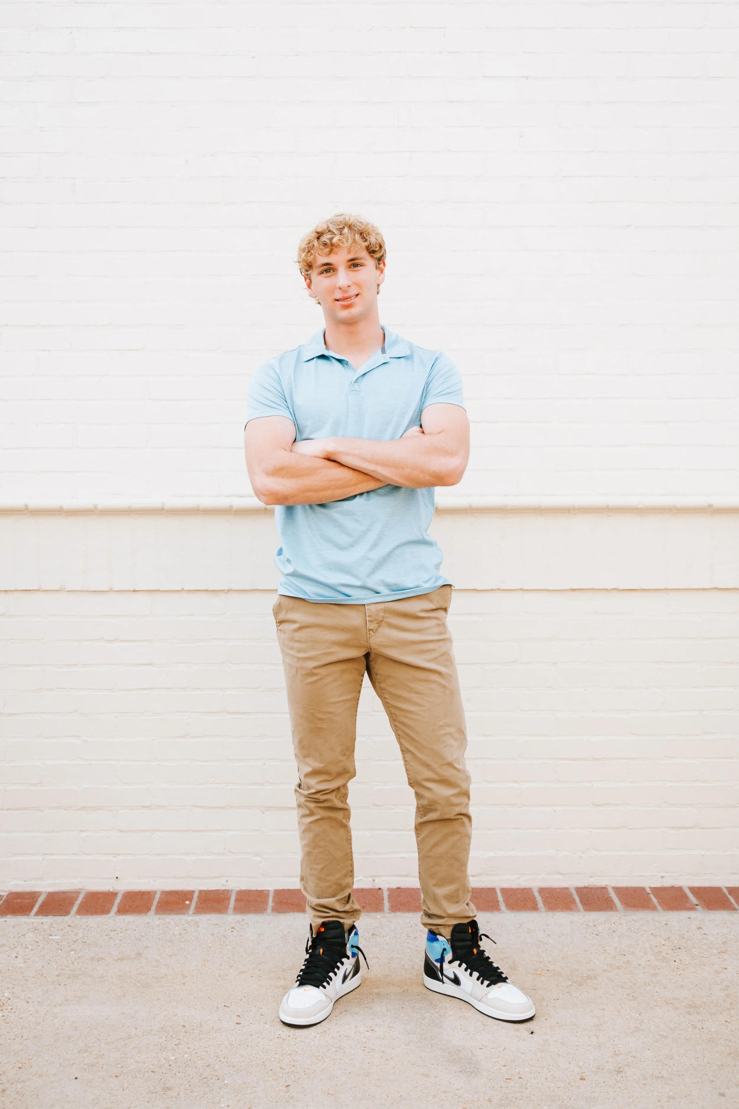 A young man with curly blonde hair standing with arms crossed in front of a white brick wall, wearing a light blue polo shirt, beige pants, and black and white sneakers.  Senior Photographer Madison, MS 