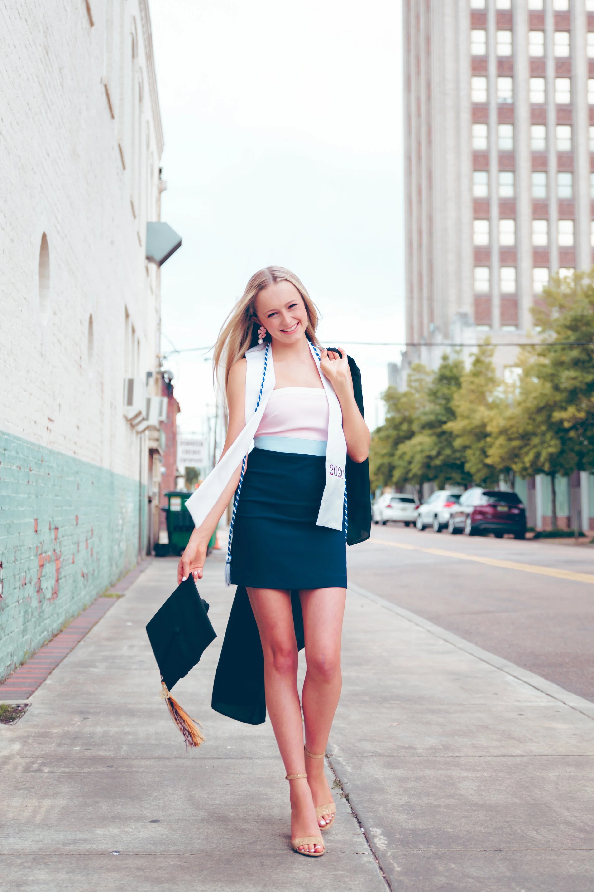 A young woman in a graduation cap and gown holding a diploma, standing on a city sidewalk with buildings and parked cars in the background, smiling and carrying her gown over her shoulder.  Senior Photographer Madison, MS 