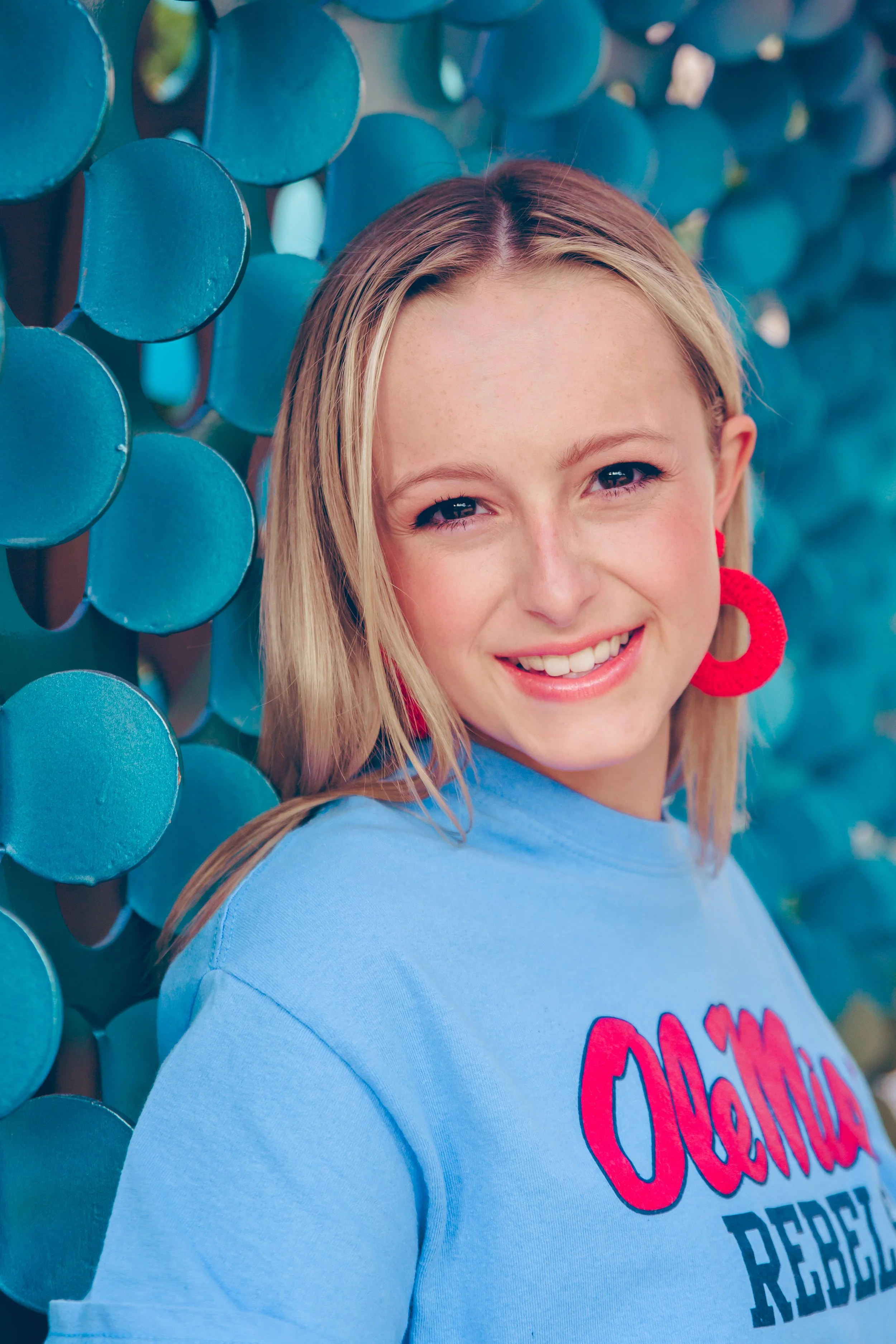 A woman with blonde hair wearing large red earrings and a light blue shirt smiling with a background of blue circular sculptures.  Senior Photographer Madison, MS 