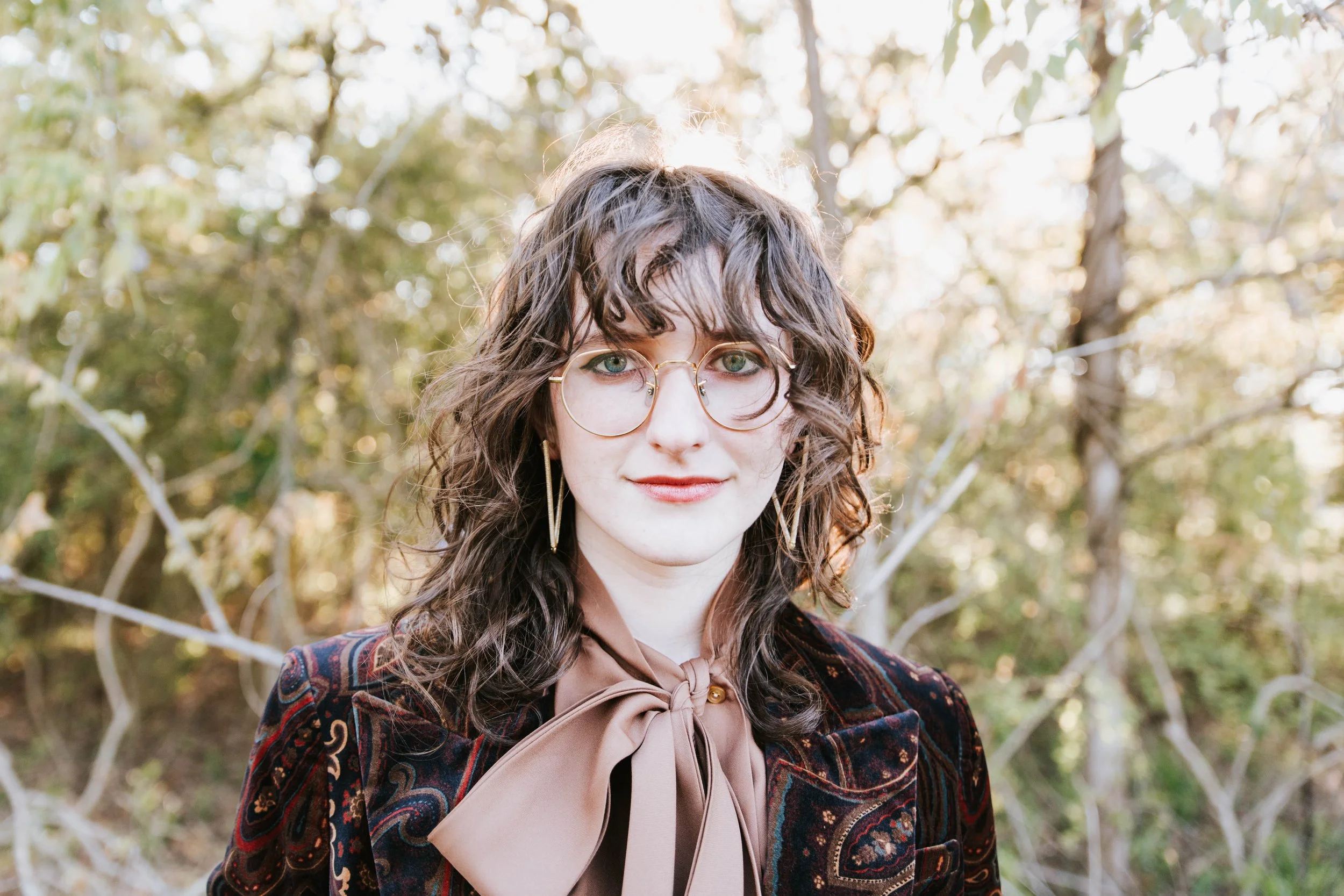 A woman with curly brown hair, glasses, and a slight smile poses outdoors with trees in the background. She wears a patterned jacket and a large beige bow tie blouse.  Senior Photographer Madison, MS 