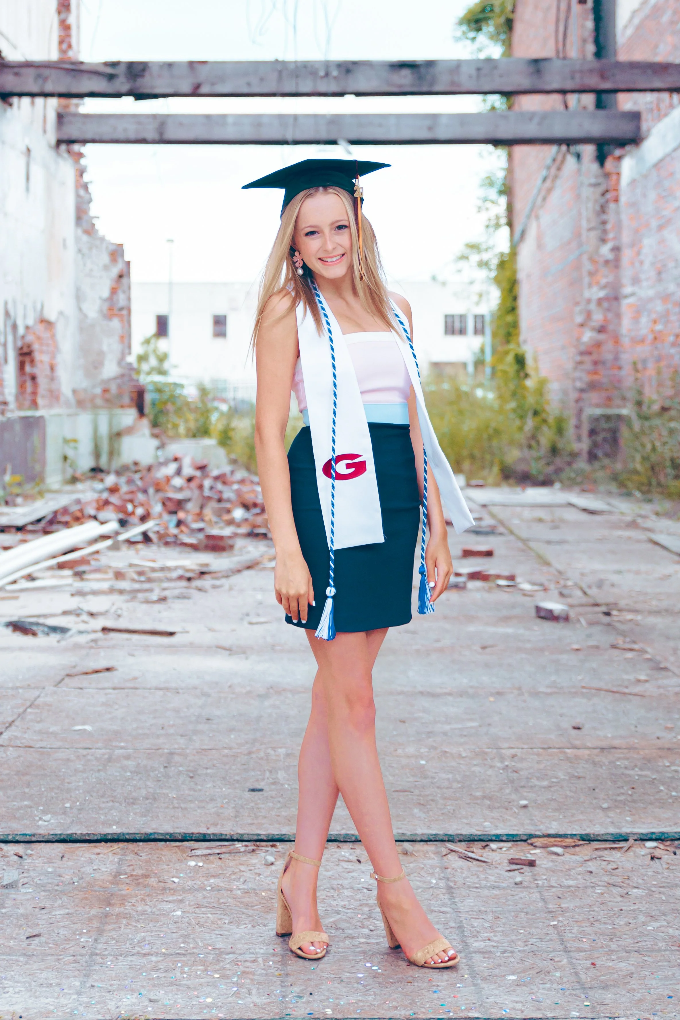 A young woman dressed in a graduate cap, gown, and sash, standing outdoors in an urban, industrial setting with exposed brick and debris.  Senior Photographer Madison, MS 