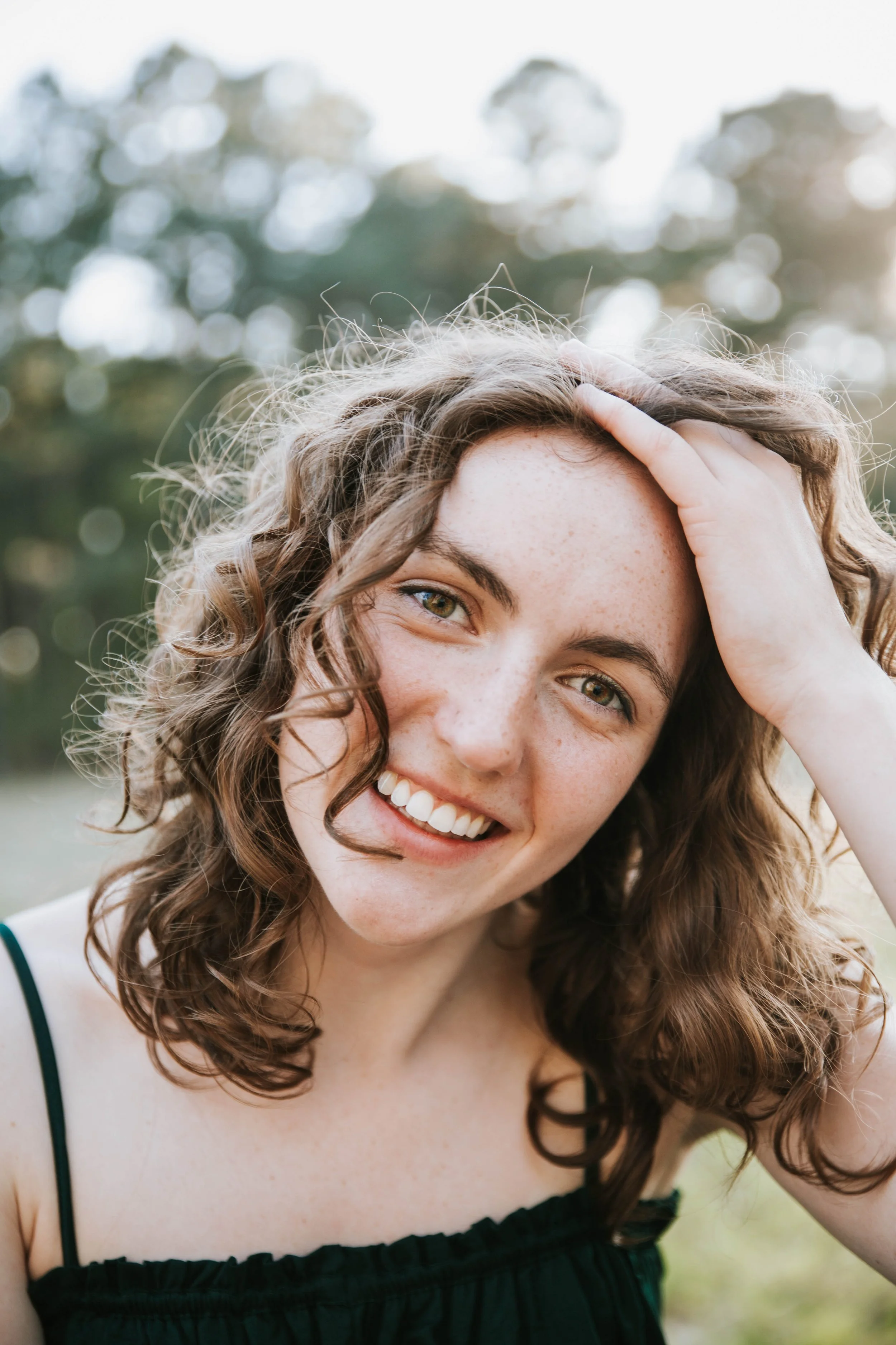 A young woman with curly brown hair and freckles smiling outdoors, holding her head with one hand.  Senior Photographer Madison, MS 