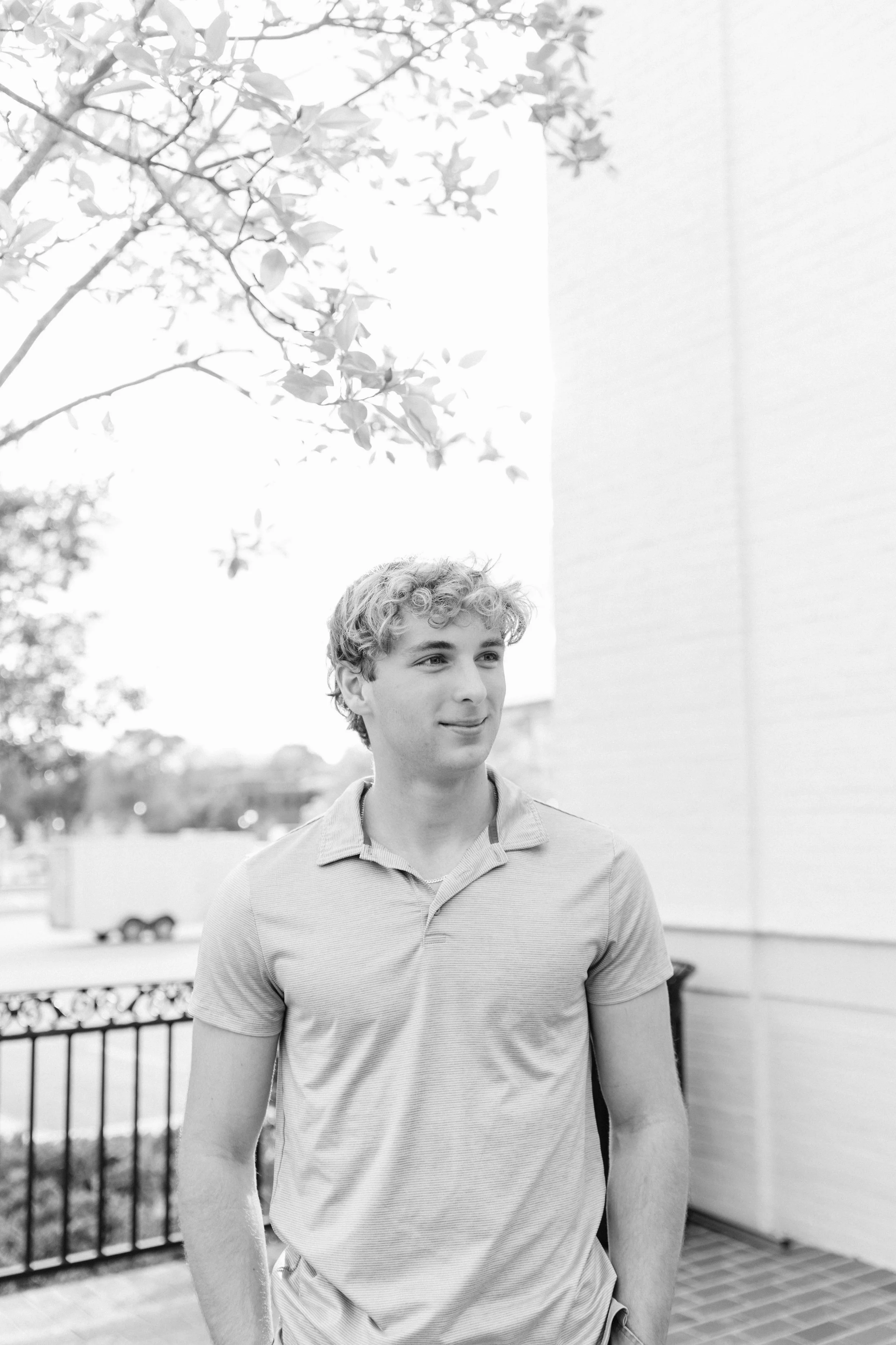 Young man with curly hair wearing a polo shirt standing outdoors near a railing and building, with trees and a parked car in the background.  Senior Photographer Madison, MS 