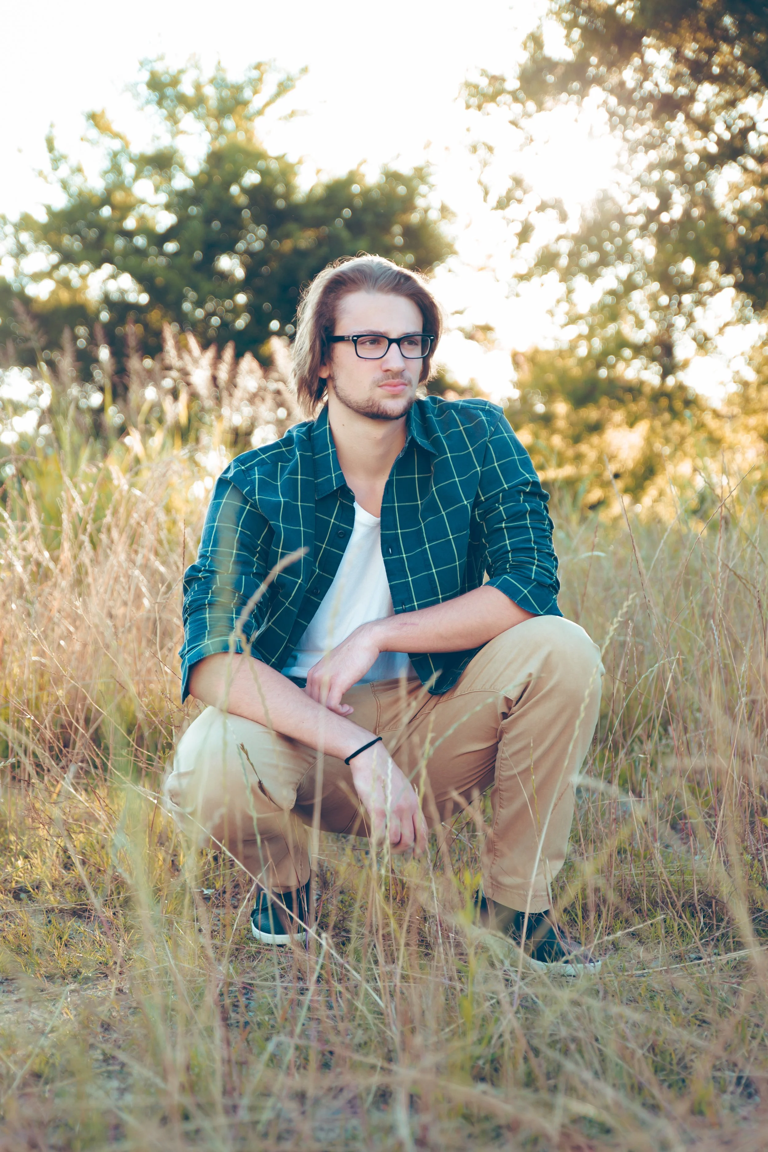 A young man with glasses, wearing a blue checkered shirt and khaki pants, squatting in tall grass in a natural outdoor setting with trees and sunlight in the background.  Senior Photographer Madison, MS 