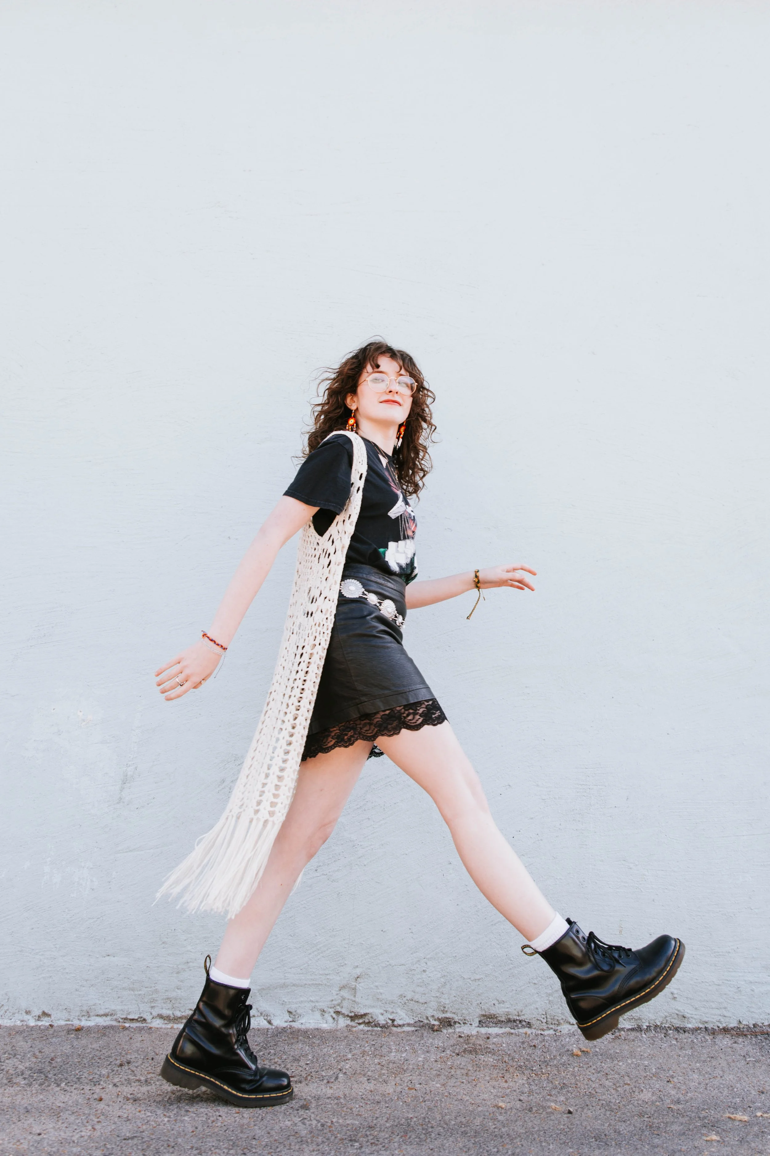 A young woman with curly brown hair, glasses, and eclectic jewelry walking in front of a plain white wall, wearing black boots, a black graphic t-shirt, a lace-trimmed skirt, and a long crochet cardigan. Senior Photographer Madison, MS 