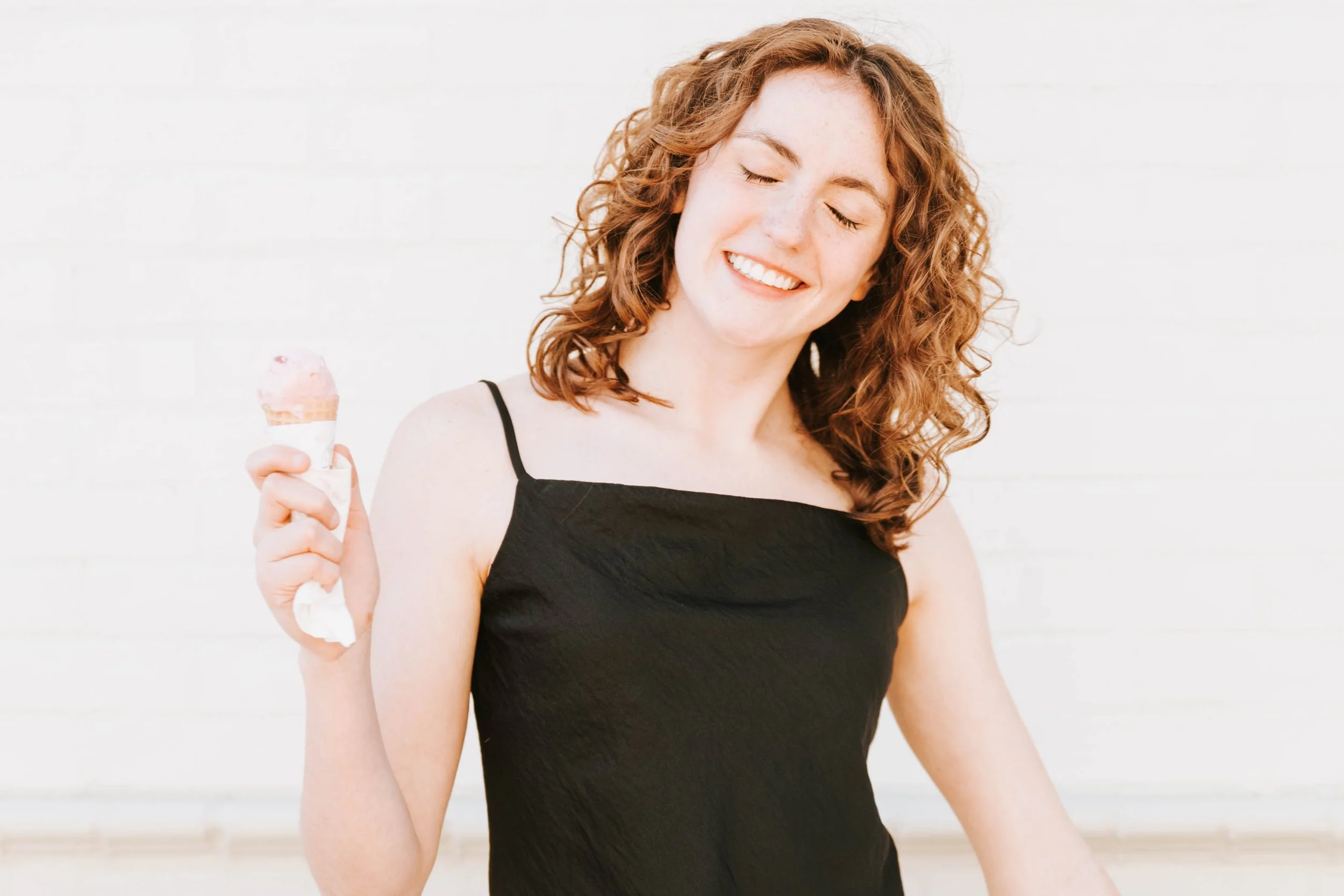 A young woman with curly red hair holding a small ice cream cone, smiling with eyes closed, wearing a black sleeveless top, standing against a white background.  Senior Photographer Madison, MS 