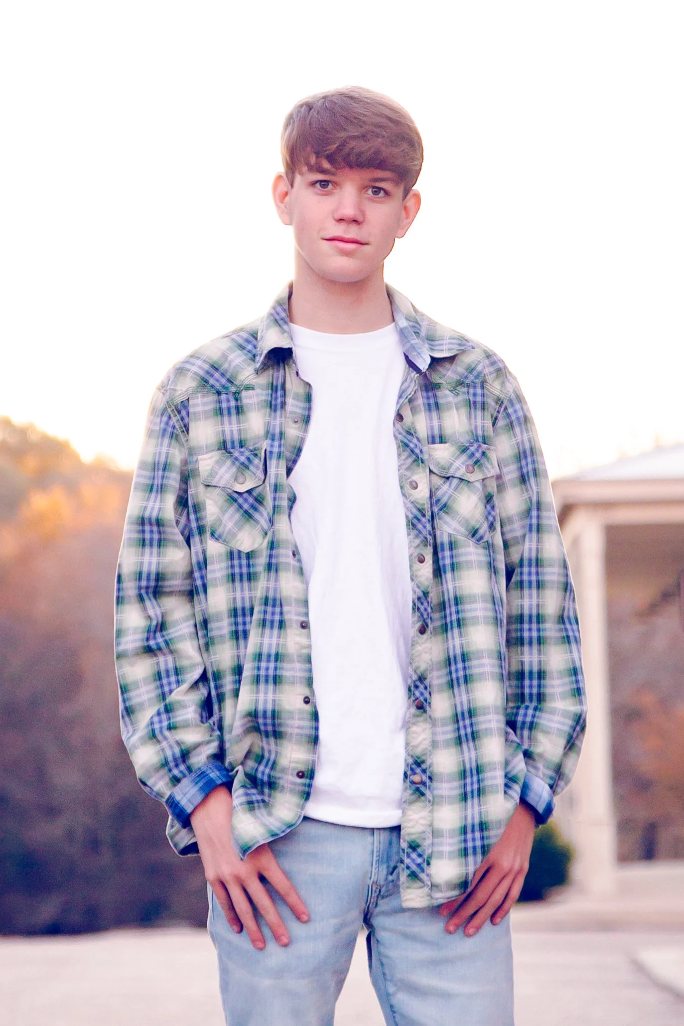 A teenage boy standing outdoors during sunset, wearing a plaid shirt over a white t-shirt and jeans, with hair styled in a side-swept manner.  Senior Photographer Madison, MS 