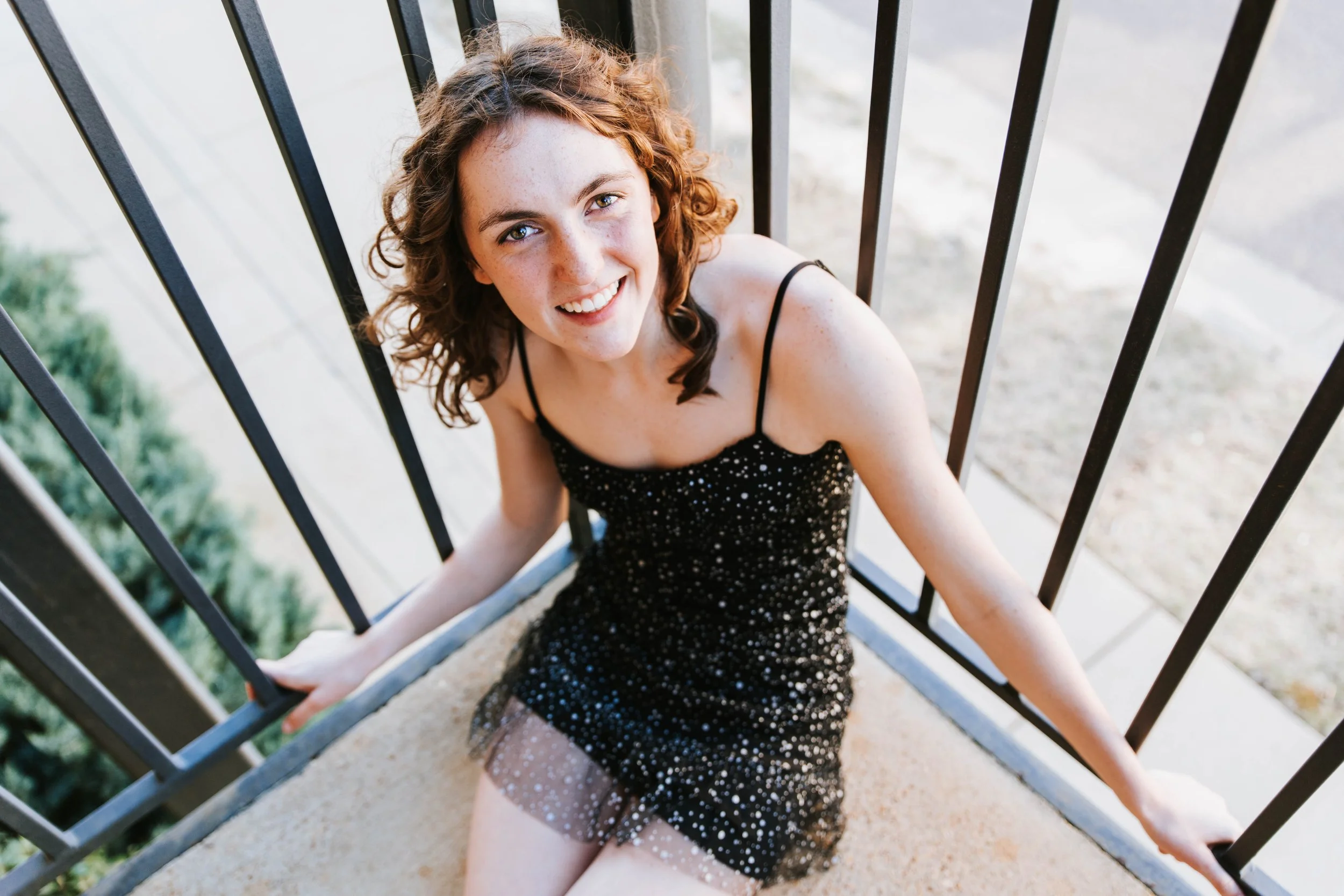 A young woman with curly brown hair and freckles, wearing a black sparkly dress with spaghetti straps, sitting on a concrete staircase and smiling at the camera.  Senior Photographer Madison, MS 