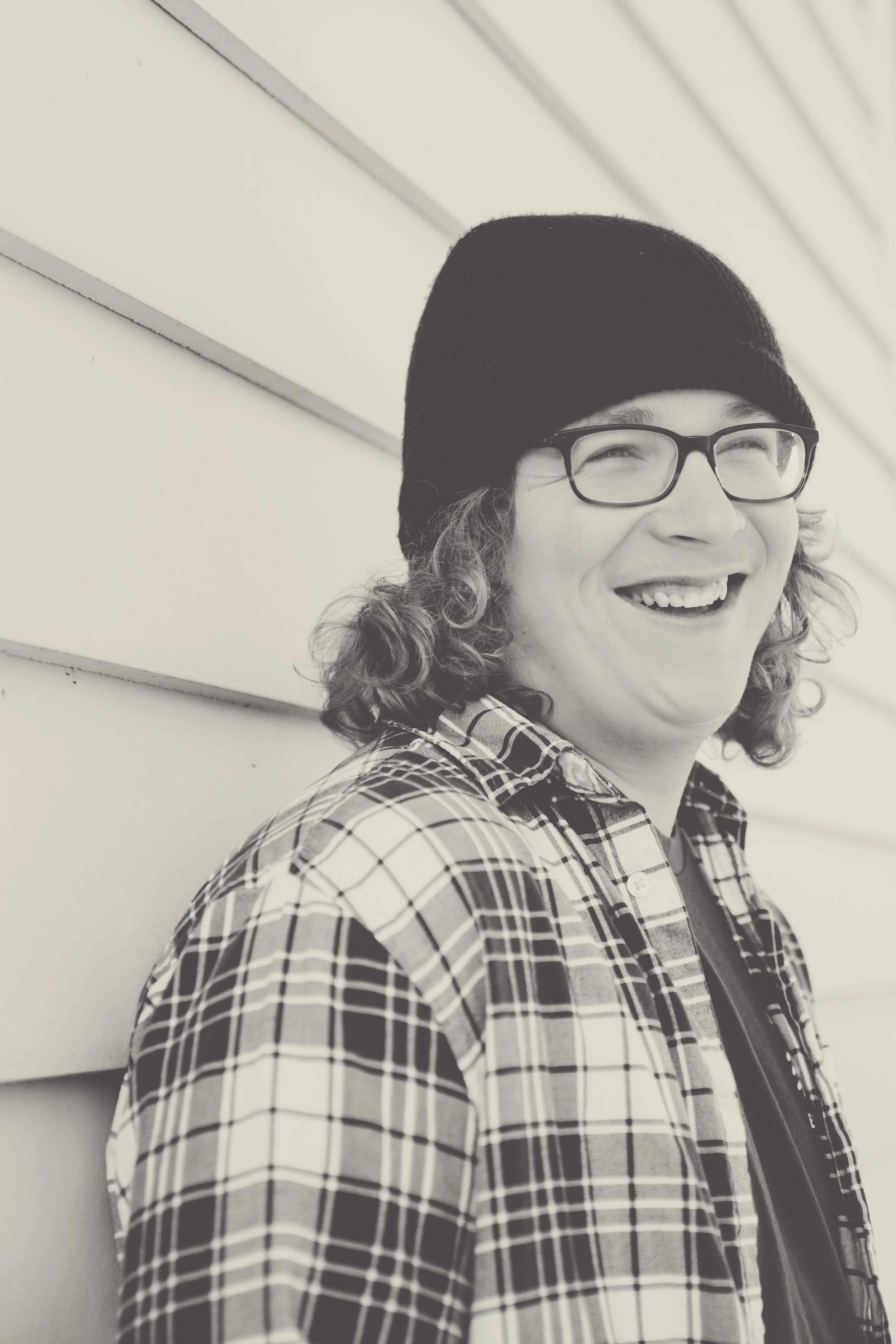 A smiling person with curly hair, glasses, and a beanie hat, wearing a plaid shirt, leaning against a wall with horizontal siding.  Senior Photographer Madison, MS 