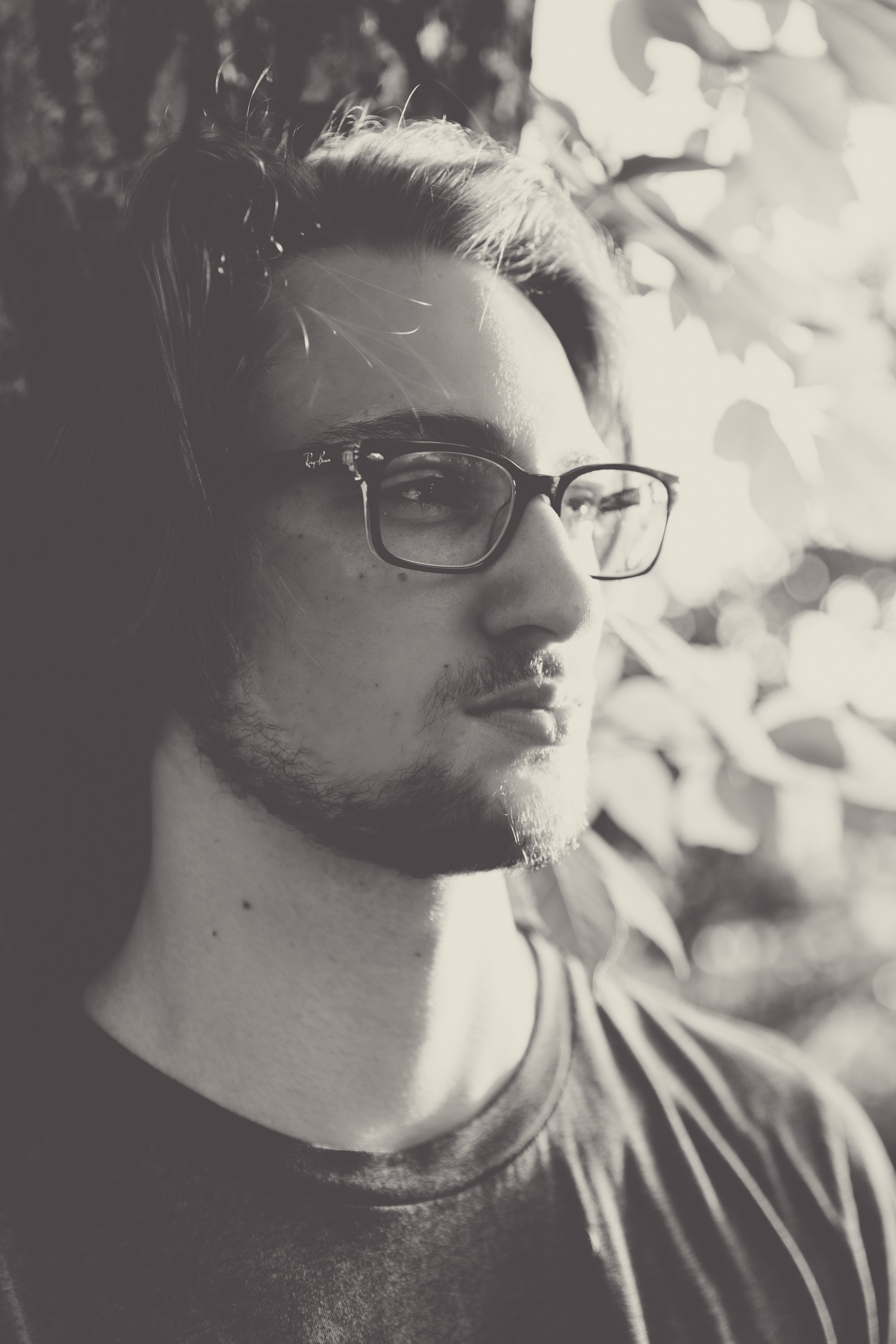 A young man with glasses, facial hair, and long hair, gazing thoughtfully into the distance outdoors with sunlight and foliage in the background.  Senior Photographer Madison, MS 