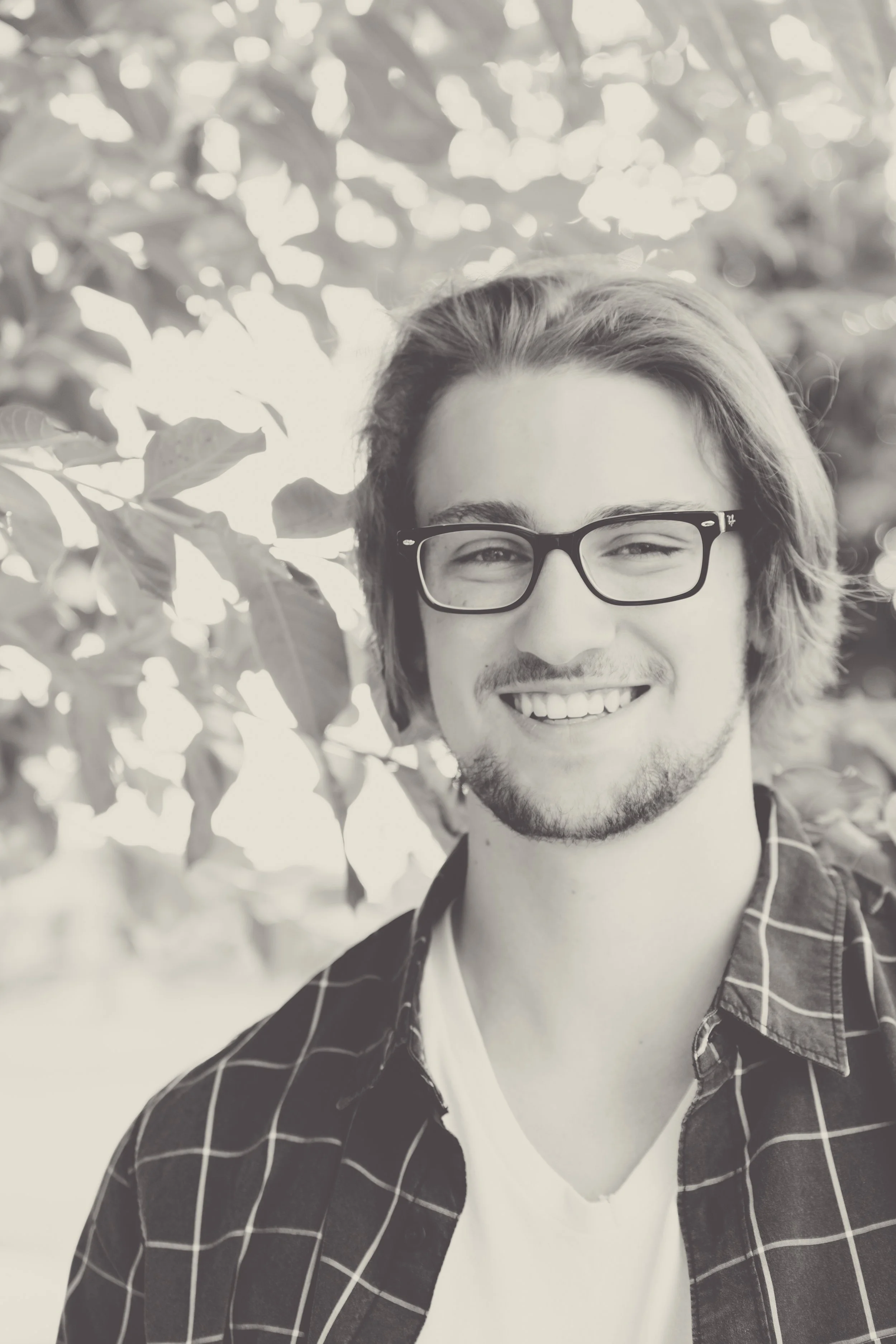 A black-and-white portrait of a young man with glasses, smiling, with shoulder-length hair, a bit of facial hair, wearing a checkered shirt and a white t-shirt, standing outdoors near leafy trees.  Senior Photographer Madison, MS 