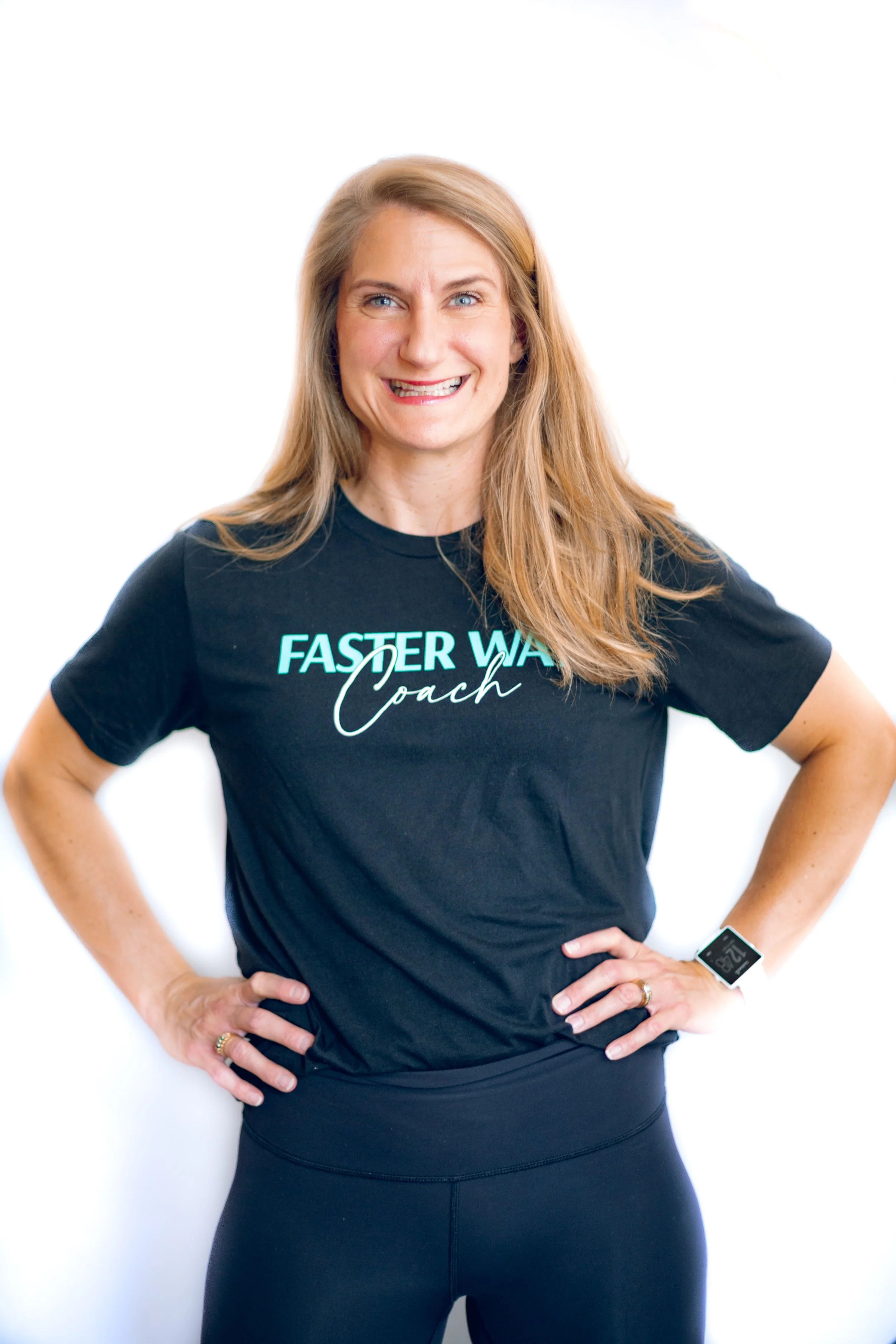 A woman with long blonde hair smiling while wearing a black 'Faster Way Coach' t-shirt and black leggings, standing against a white background.