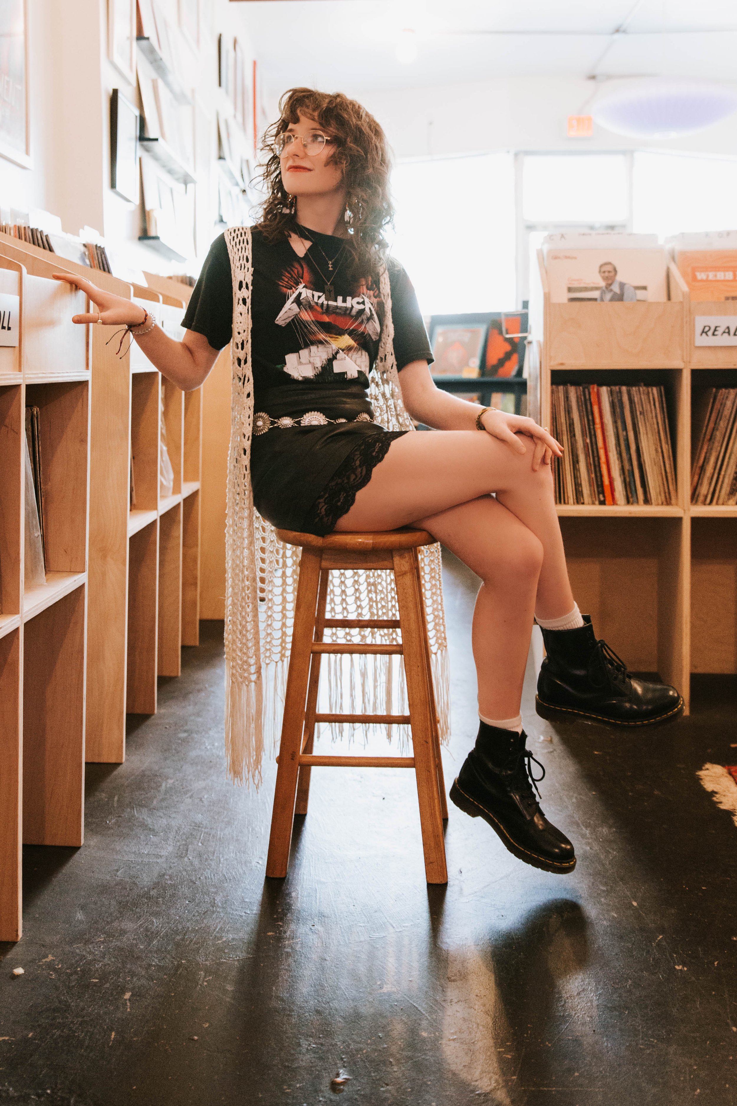 Young woman with curly hair and glasses sitting on a wooden stool in a record store, wearing a Guns N' Roses t-shirt, black lace skirt, and combat boots, surrounded by shelves of vinyl records. Senior Photographer Madison, MS 
