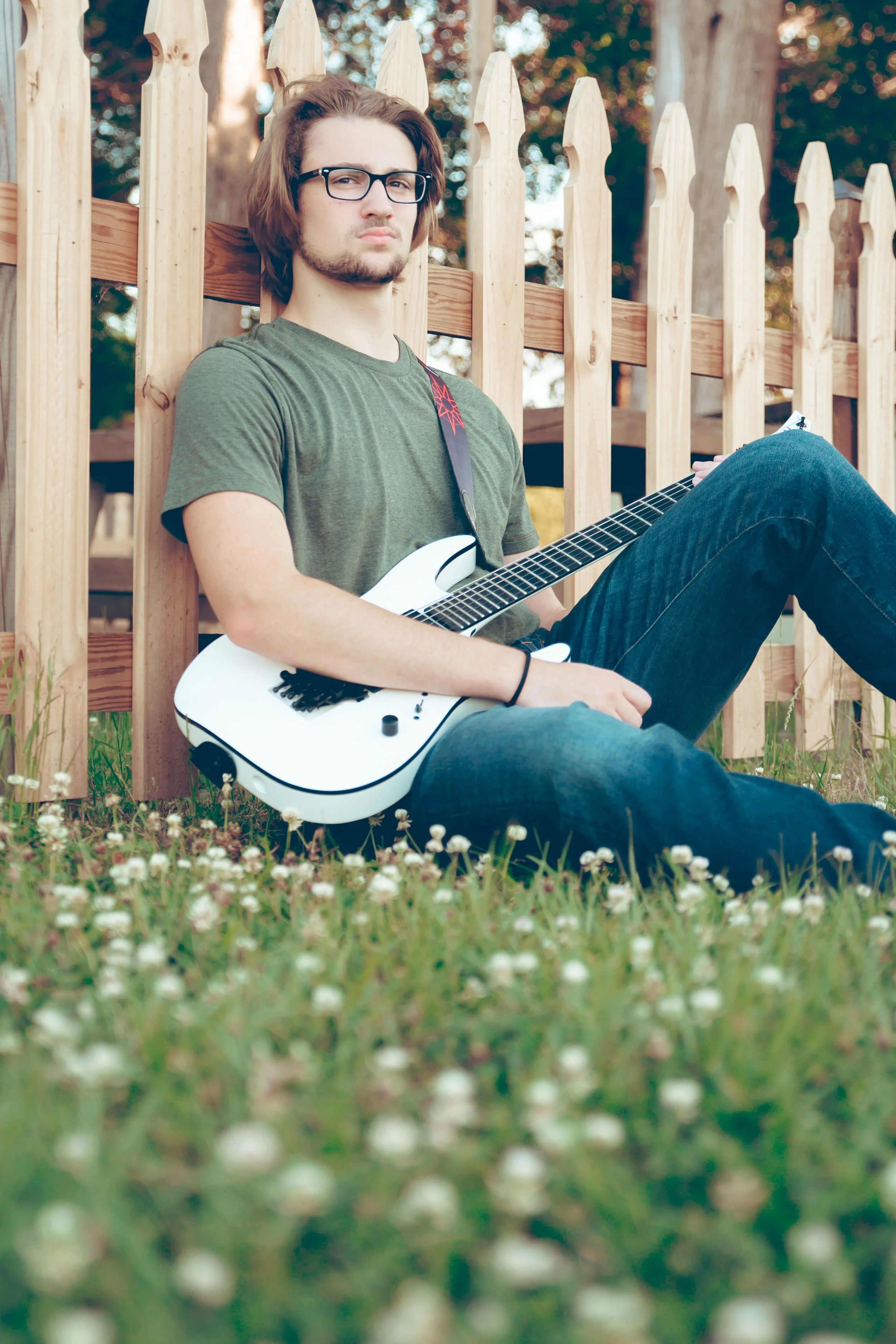 Young man sitting on grass with white electric guitar, leaning against a wooden fence, outdoors on a sunny day.  Senior Photographer Madison, MS 