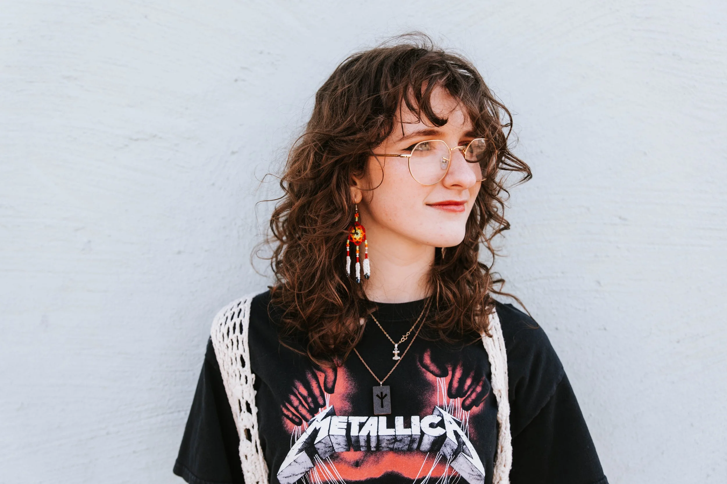 A woman with curly brown hair, wearing glasses, a Metallica t-shirt, and colorful earrings, standing against a white wall.  Senior Photographer Madison, MS 