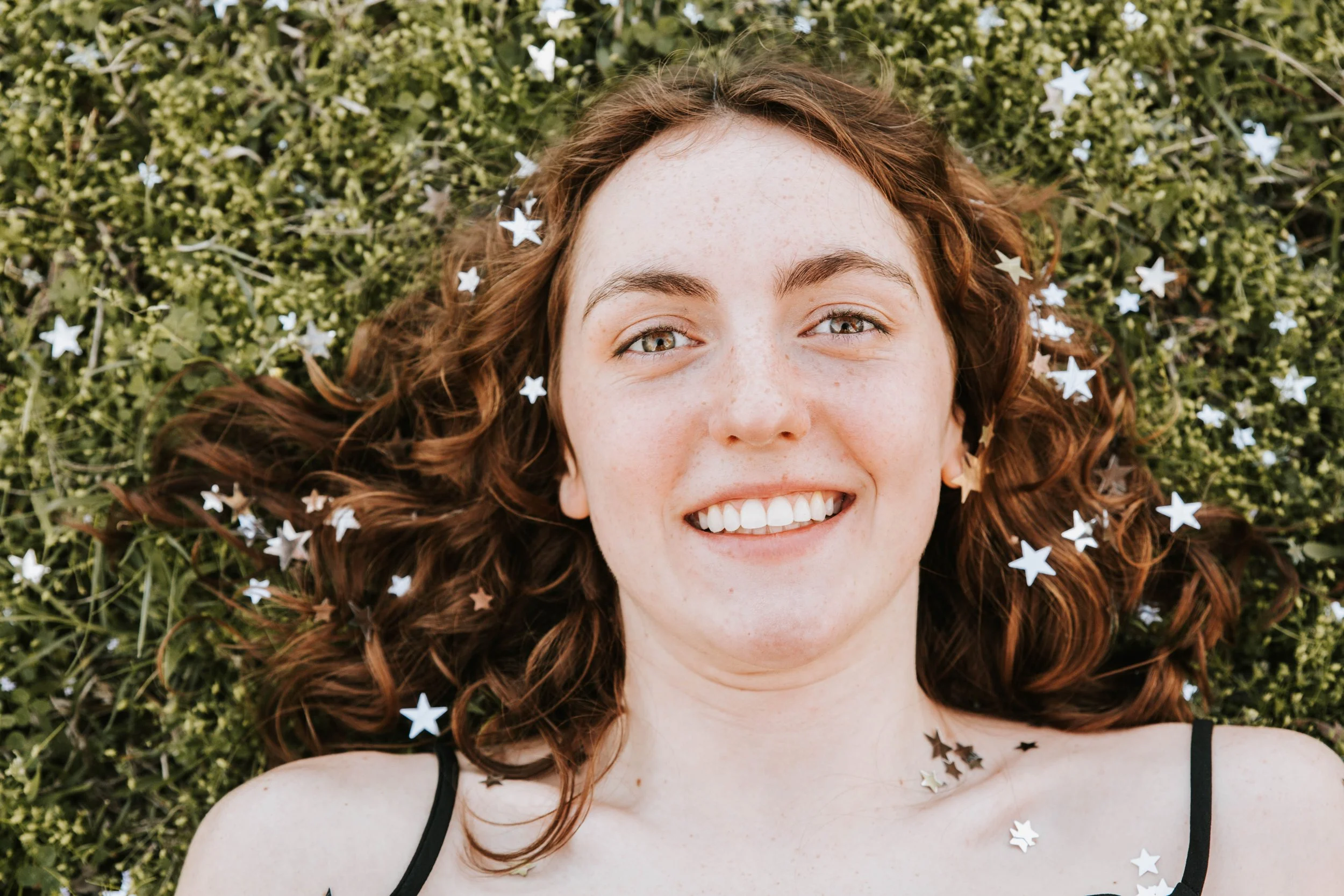 A woman with curly hair lying on grass with star-shaped confetti around her. Senior Photographer Madison, MS 