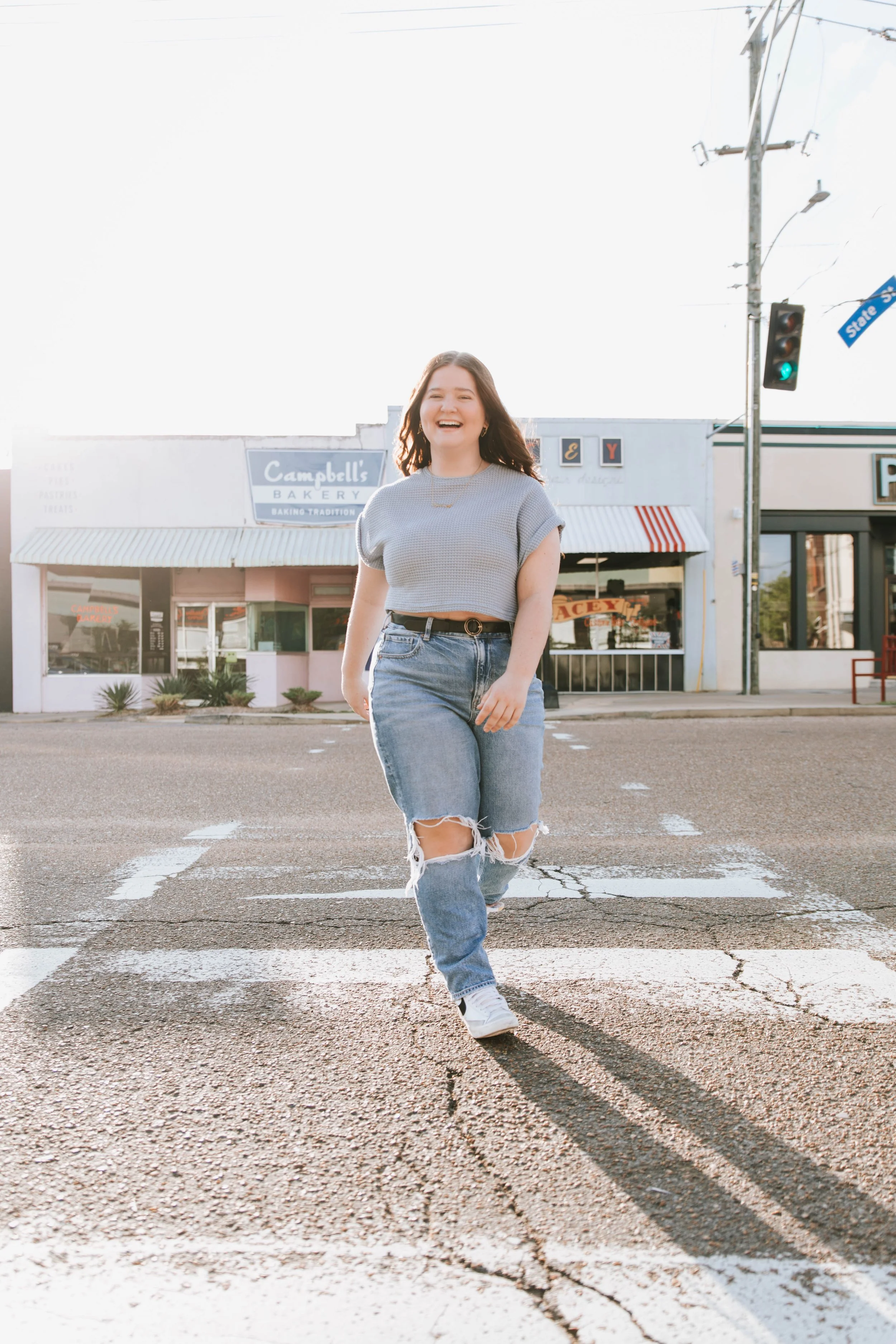 A young woman walking across a crosswalk, smiling, with a bakery storefront and street sign in the background.  Senior Photographer Madison, MS 