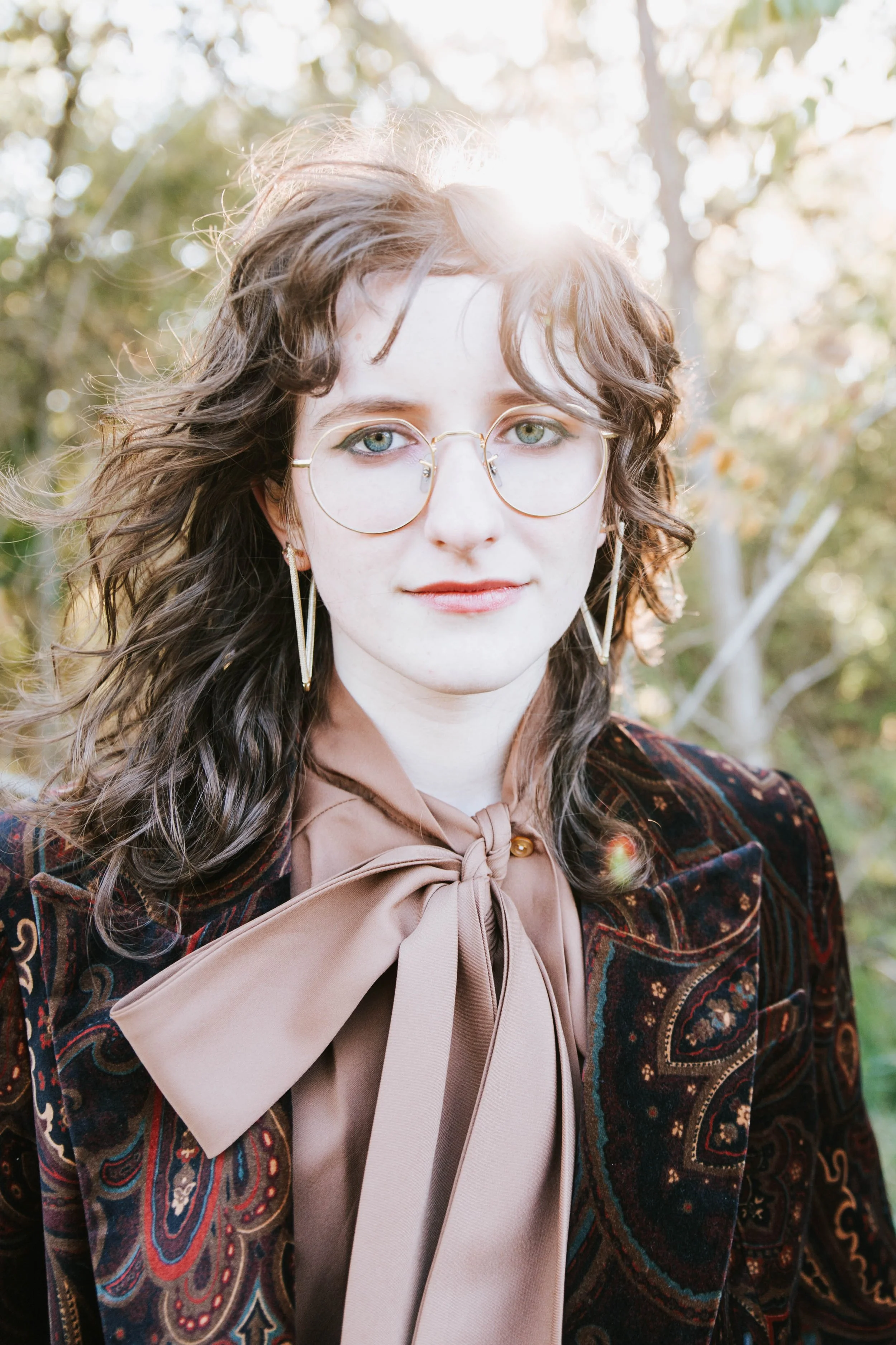 A woman with wavy brown hair and glasses standing outdoors with sunlight behind her, wearing earrings, a patterned jacket, and a beige blouse with a bow tie.  Senior Photographer Madison, MS 