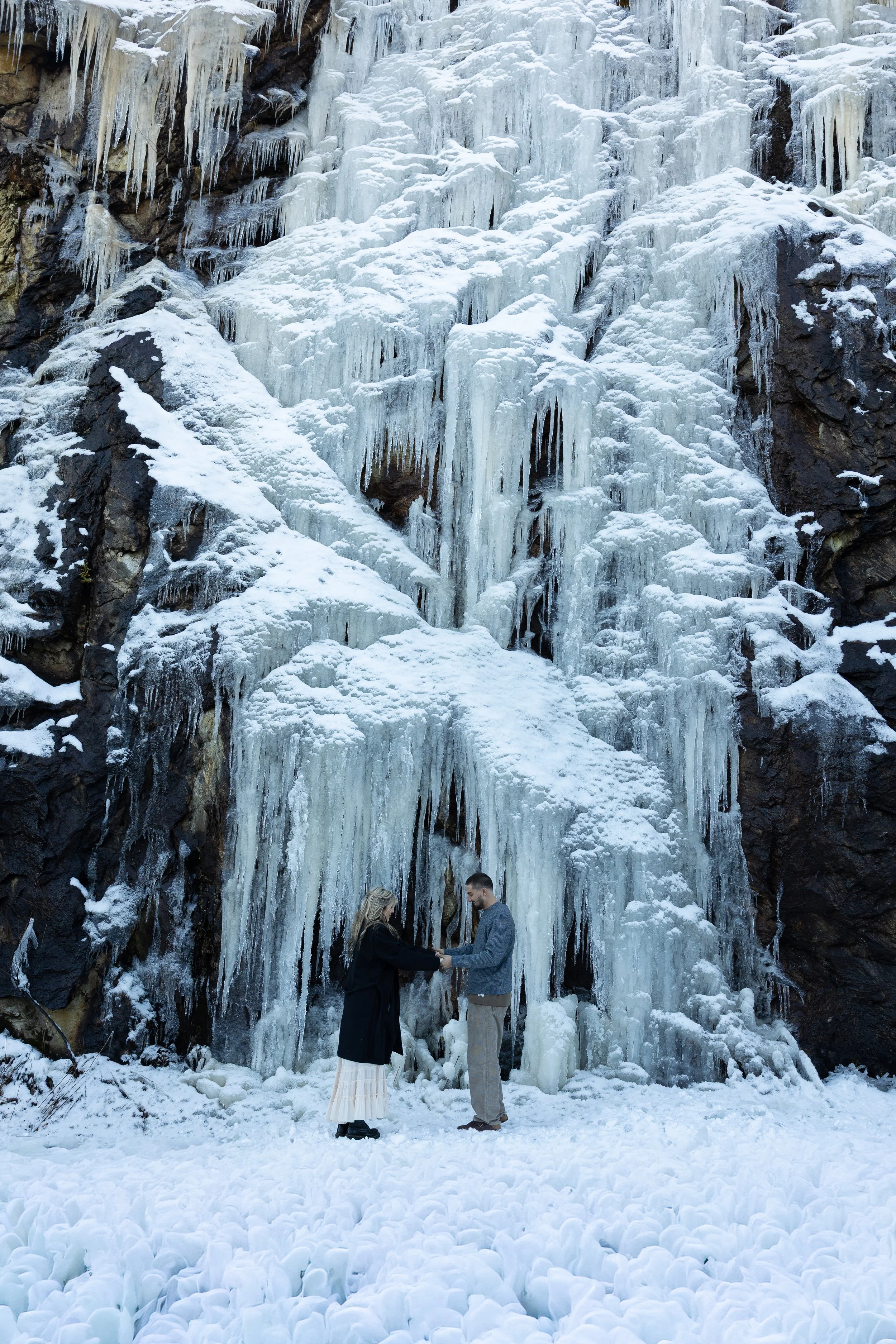 A couple standing and holding hands in front of a large, frozen waterfall with icicles and snow, in a winter landscape.