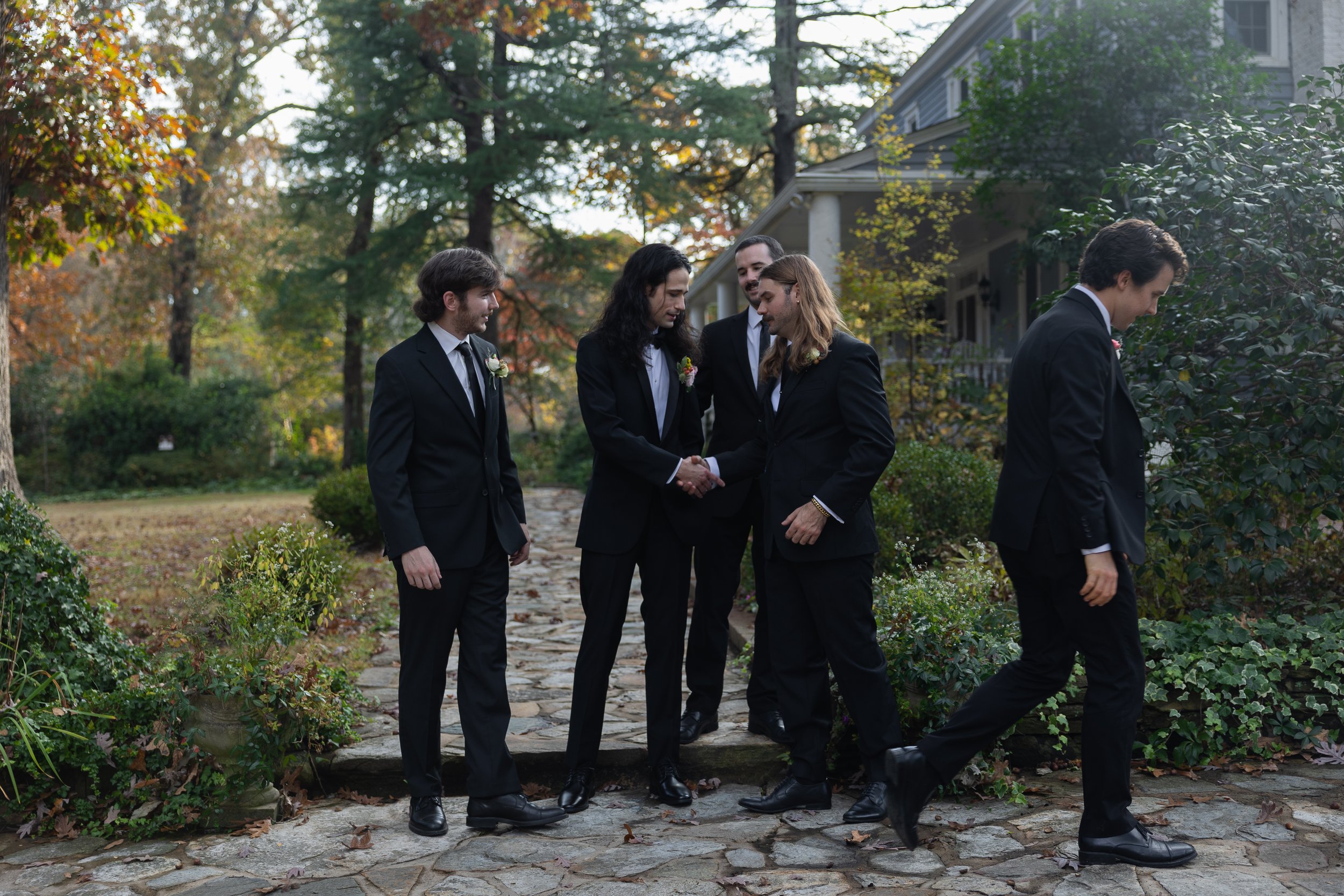 Group of five men in formal black suits and ties standing and walking outdoors in a garden, with trees and a house in the background, during fall.