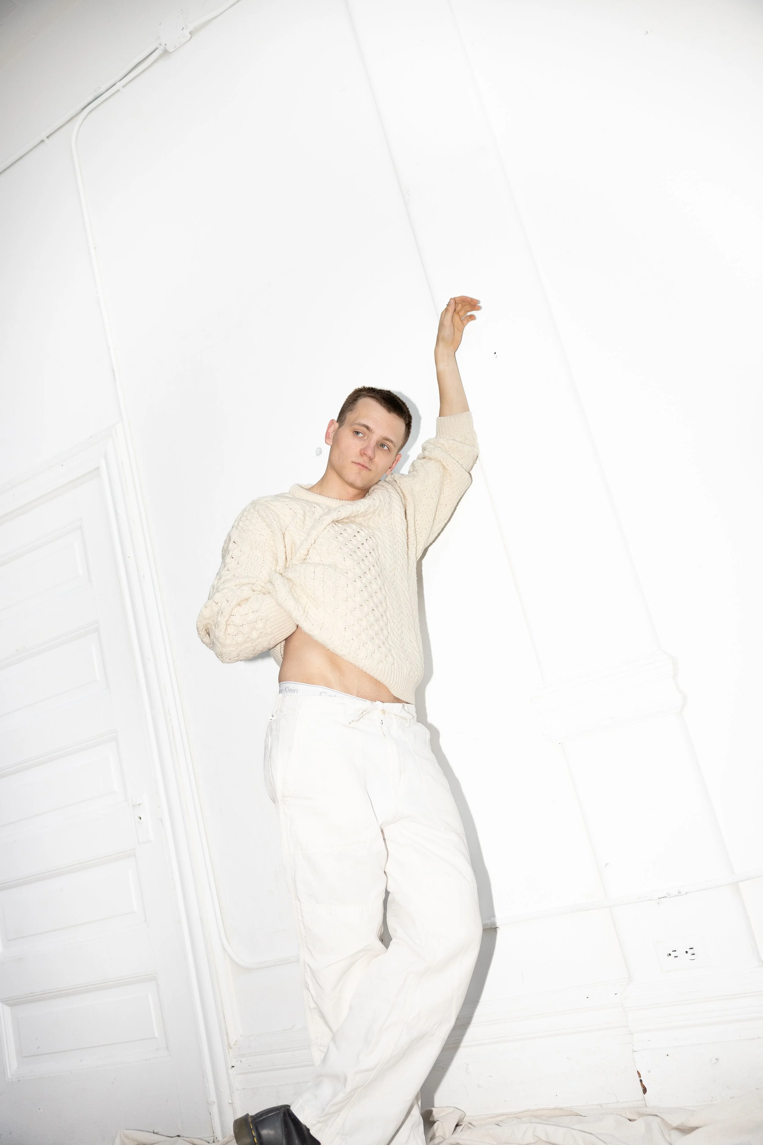 Young man standing against a white wall with one arm raised and wearing a beige sweater and white pants.