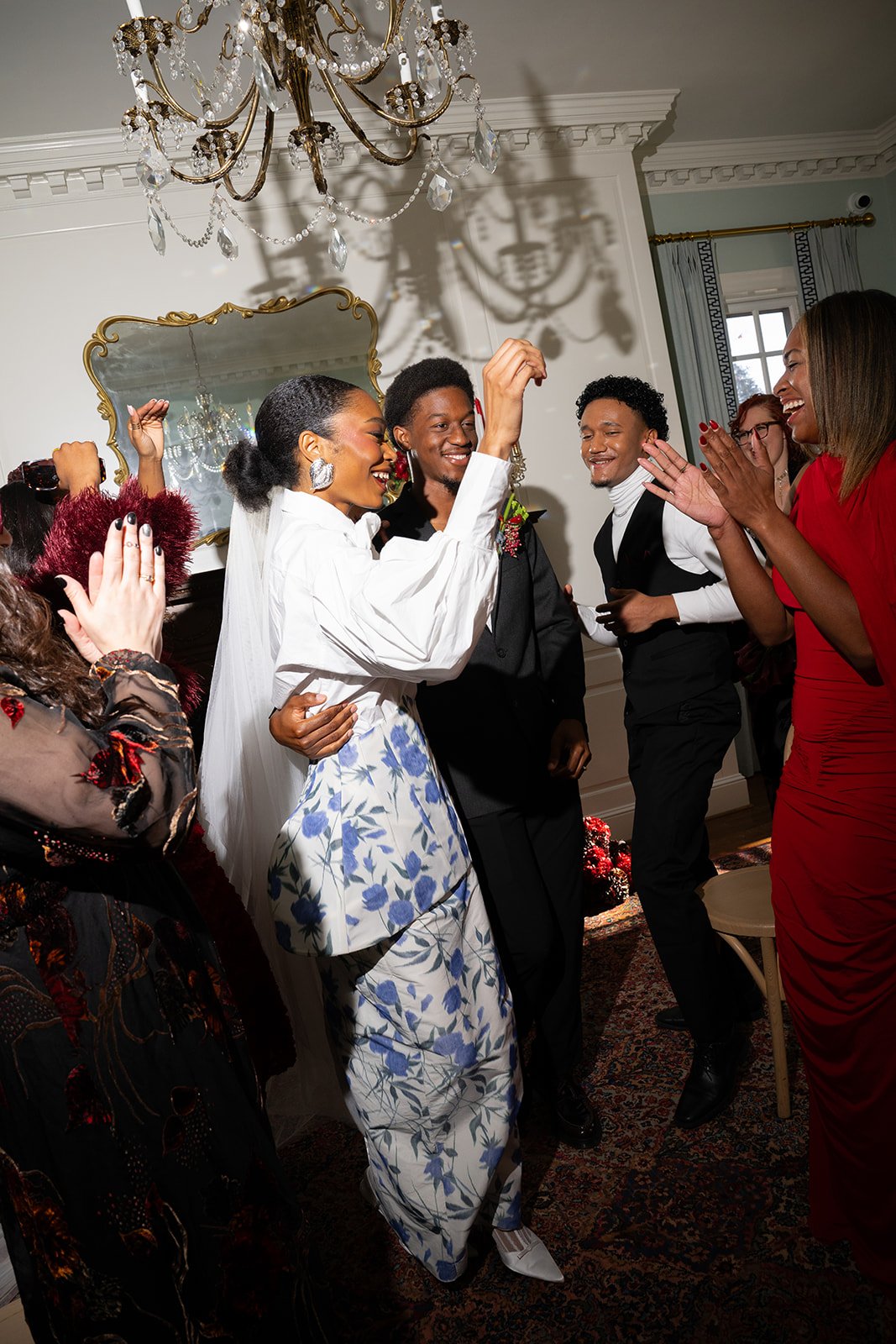 People celebrating at a wedding reception in an elegant room with chandelier and mirror, including a bride in white and a groom in gray suit surrounded by friends and family clapping and smiling.