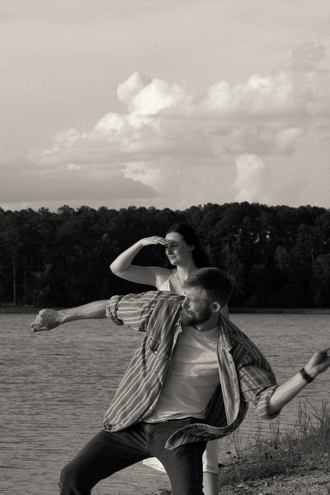 A black and white photo of a man and woman by a lake, with the man striking a pose and the woman looking into the distance and shielding her eyes with her hand, during the evening or sunset.
