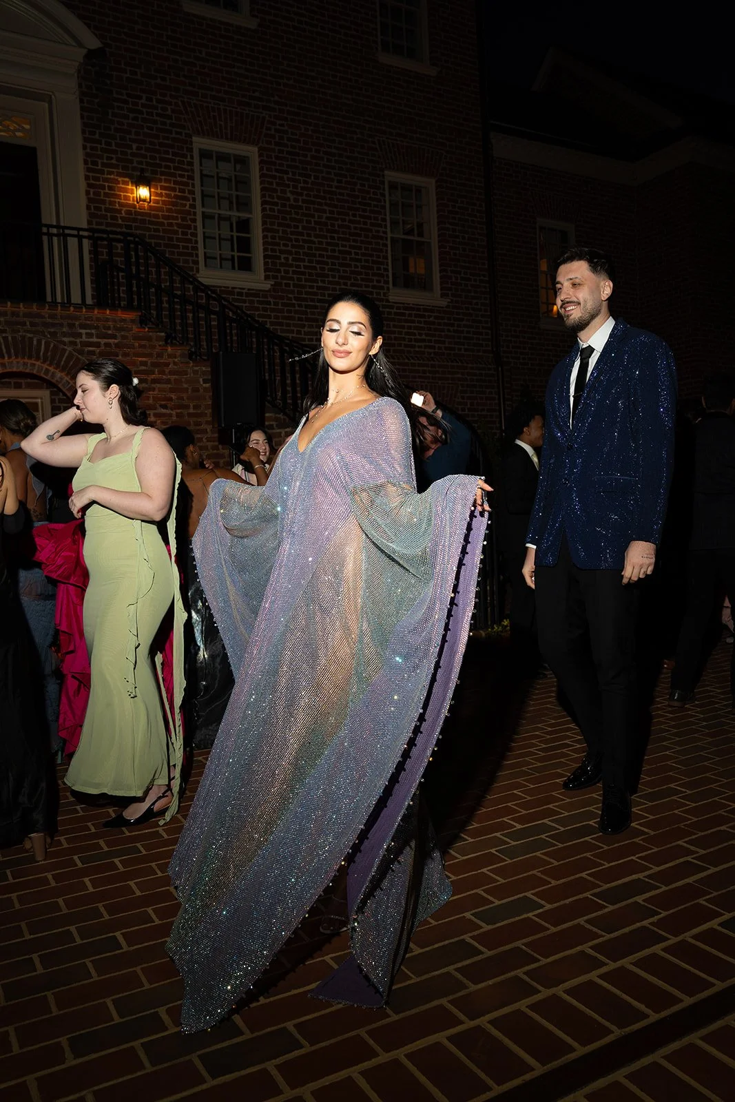 A woman in a shimmering, flowing gown with batwing sleeves walking at a formal evening event, accompanied by a man in a dark, glittery blazer and black pants.
