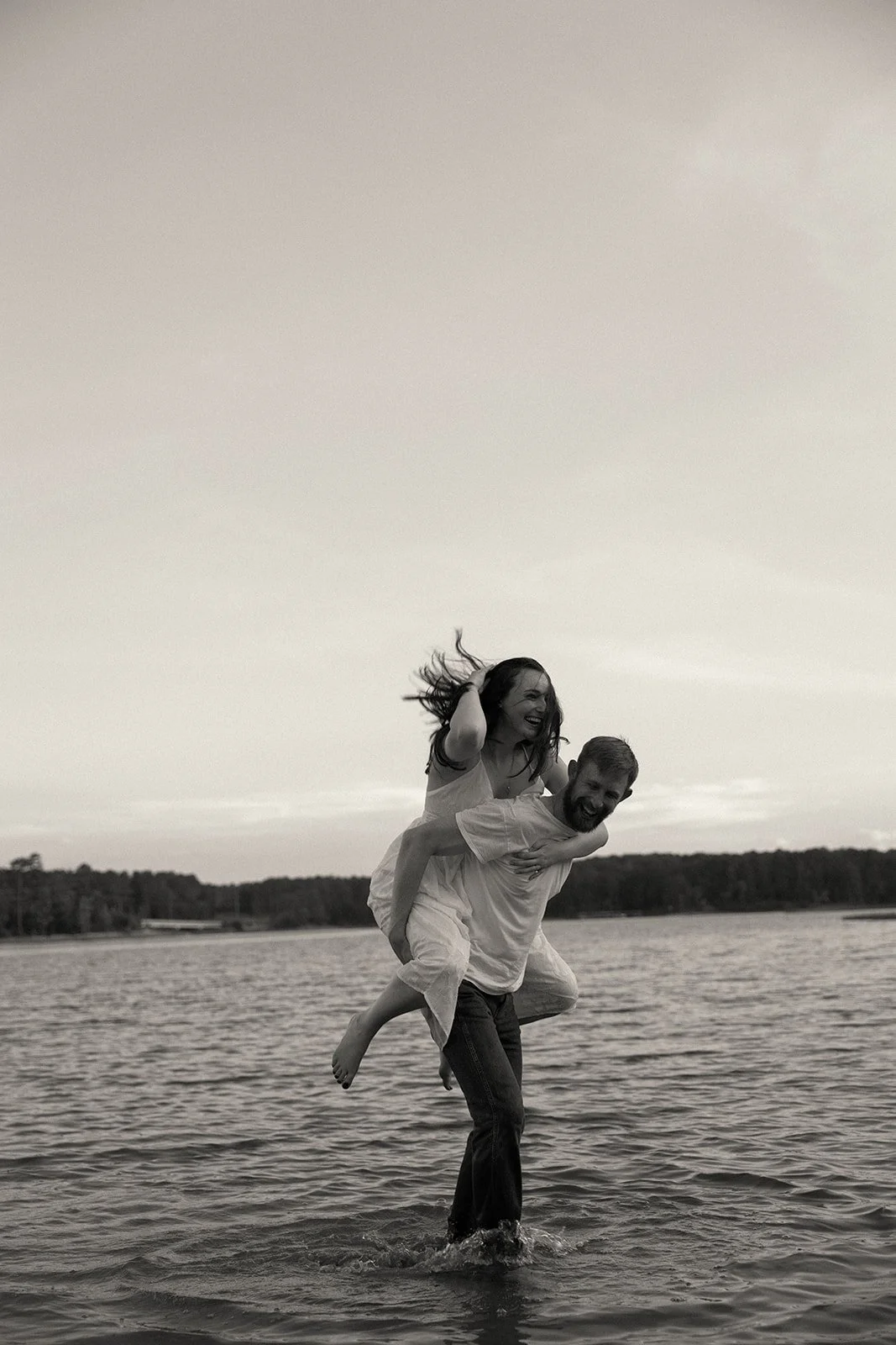 A man giving a woman a piggyback ride in a lake, both smiling and enjoying themselves, with the woman holding her hair back, in black and white.