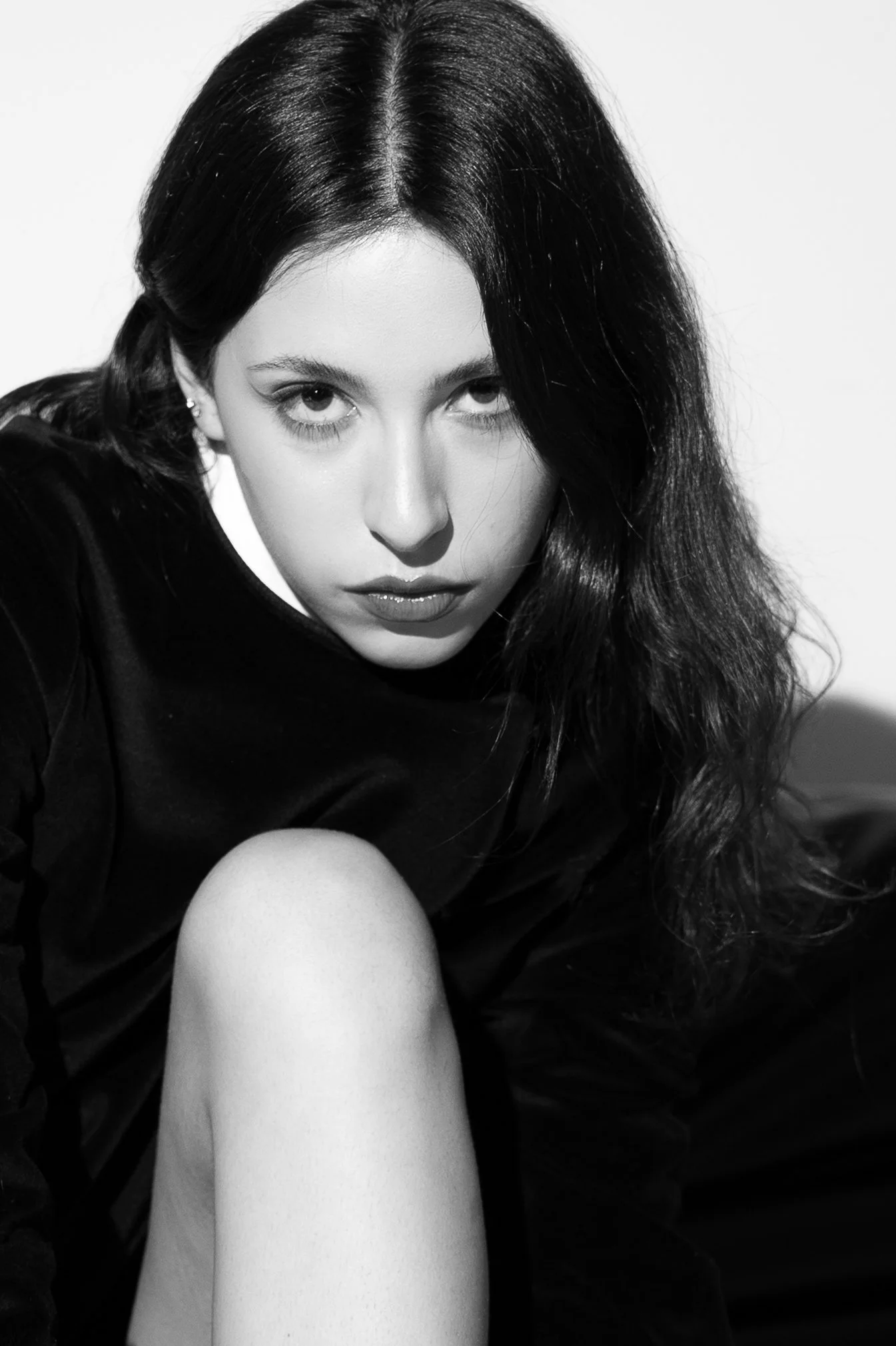 A black and white portrait of a young woman with long dark hair, intense gaze, wearing a dark top, with her knee visible in the foreground.