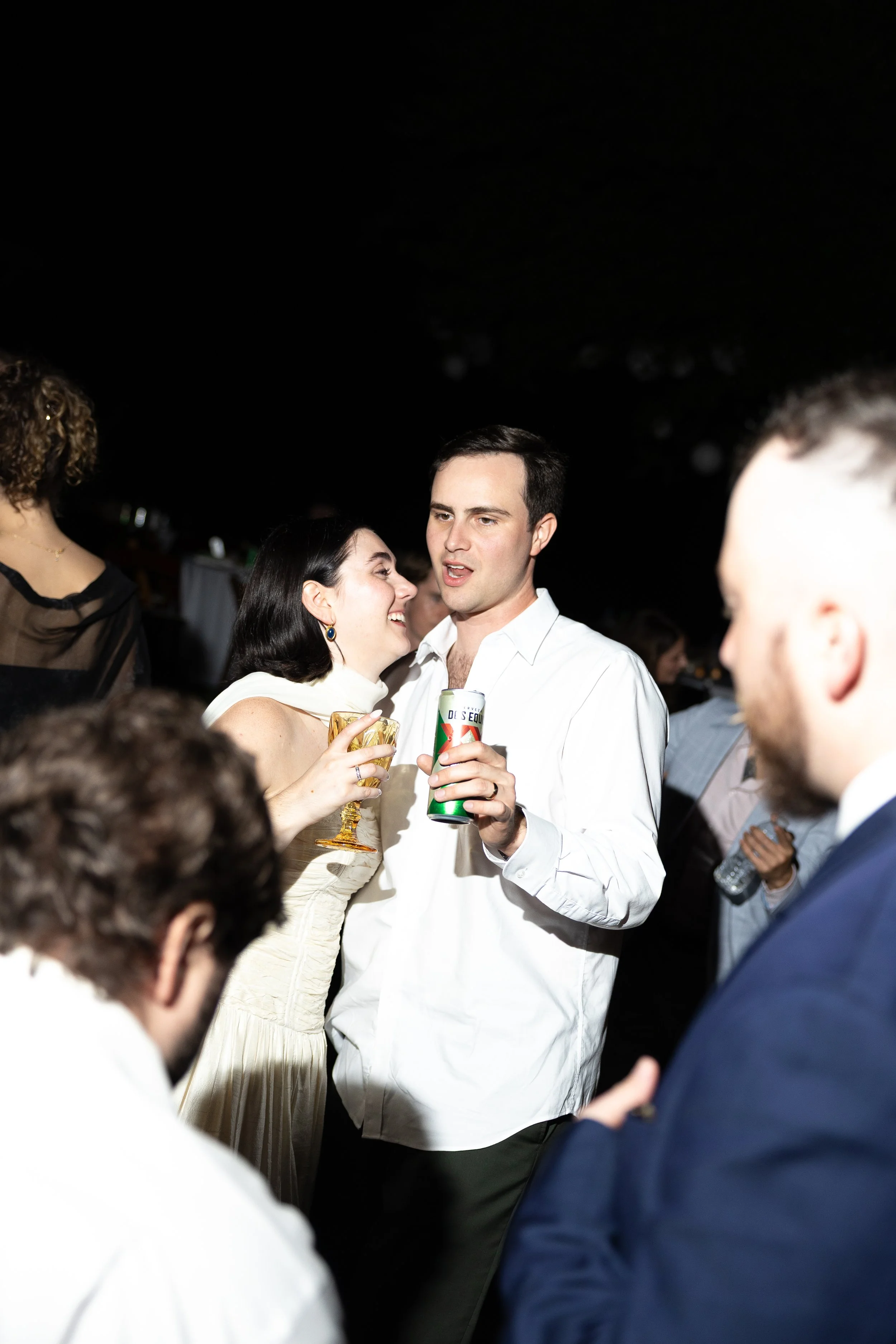 People socializing at a party, two individuals in the center holding drinks and engaging in conversation, dim background with other guests.