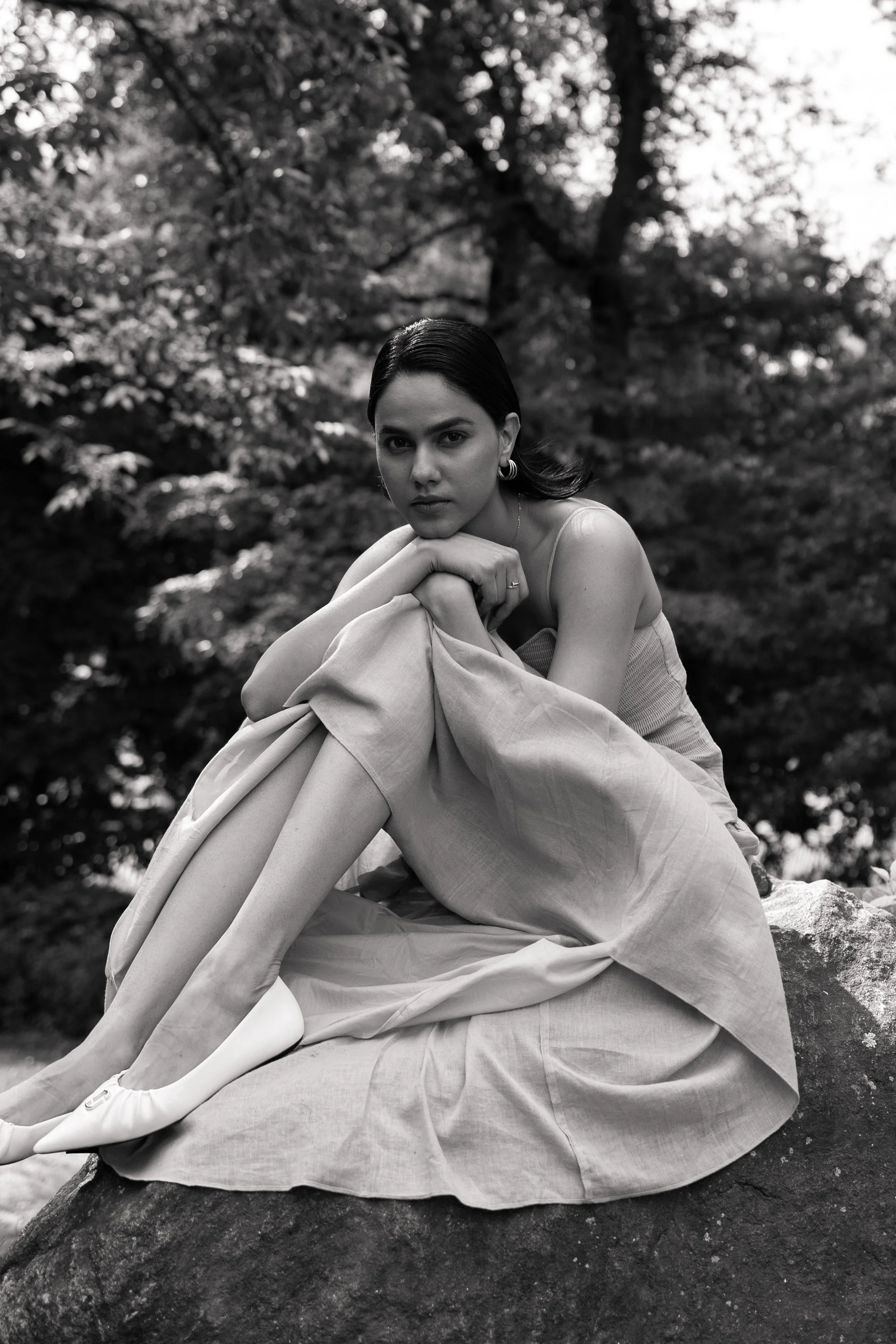 Black and white photo of a young woman with dark hair sitting outdoors on a rock, with her chin resting on her crossed arms, surrounded by trees with leaves and branches in the background.