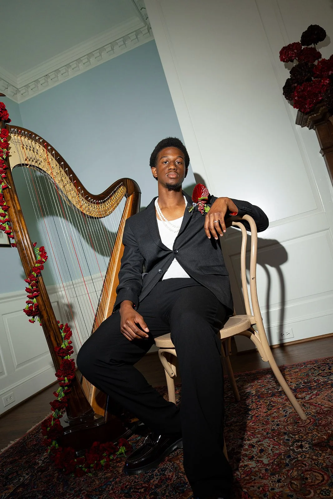 A young man in a black suit with a white shirt and pearl necklace sitting on a chair in front of a harp decorated with red flowers in a well-lit room with white walls and wooden floors.