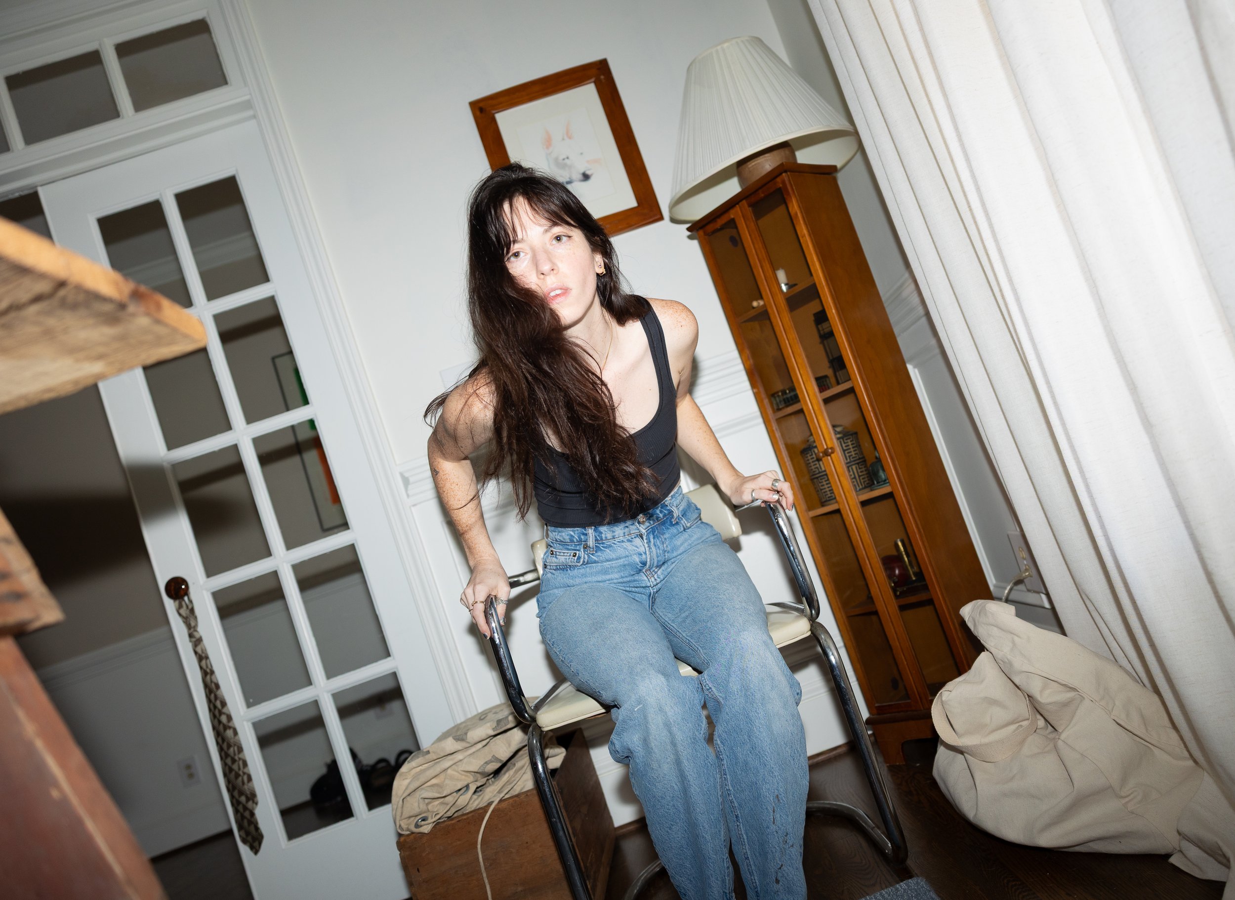 Woman with long dark hair wearing a black tank top and blue jeans sitting on a chair in a room with white walls, a wooden cabinet, and curtains.