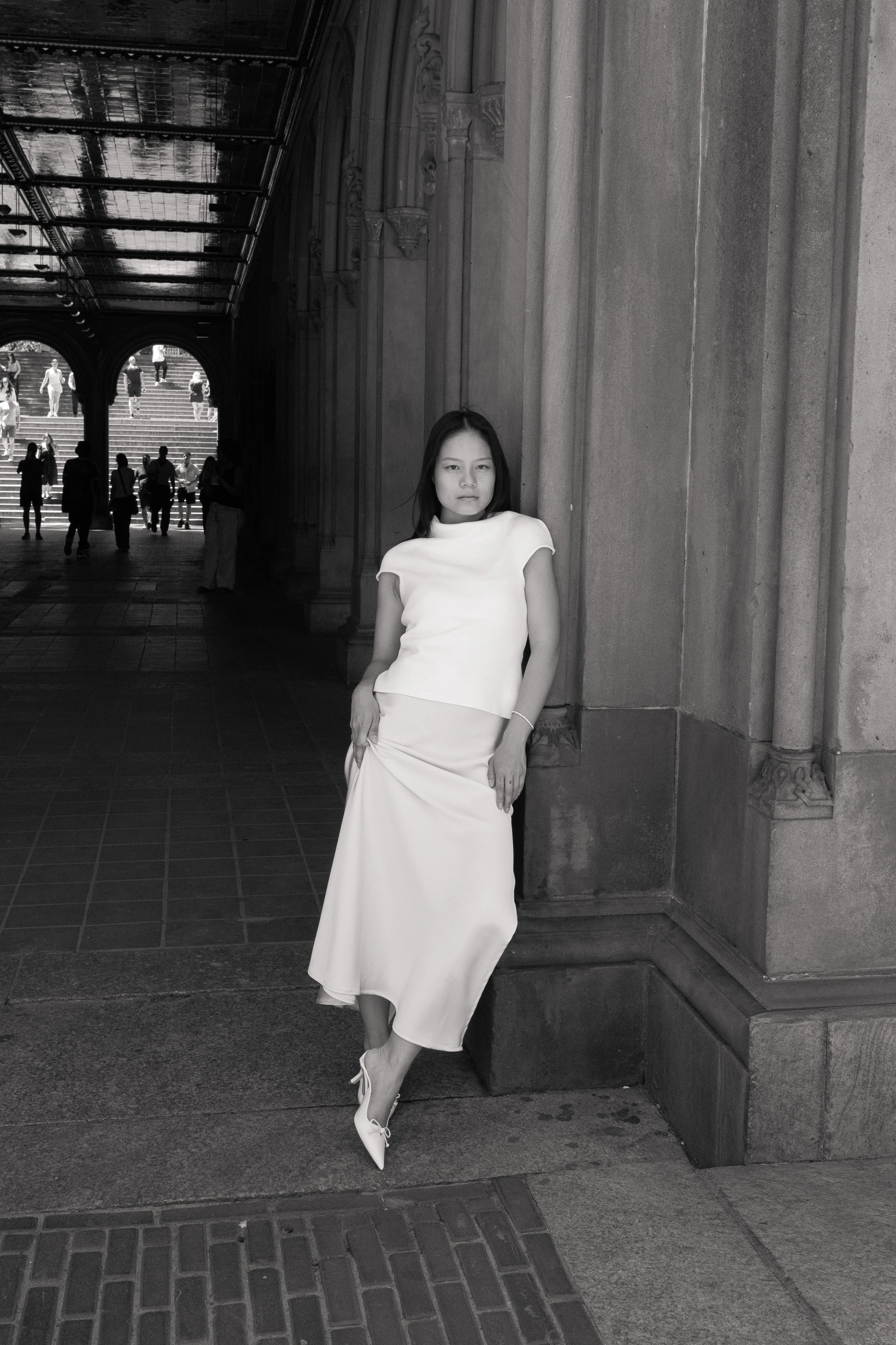 A woman in a white dress and high heels leaning against a stone column inside a building, with a large window and other people walking in the background.