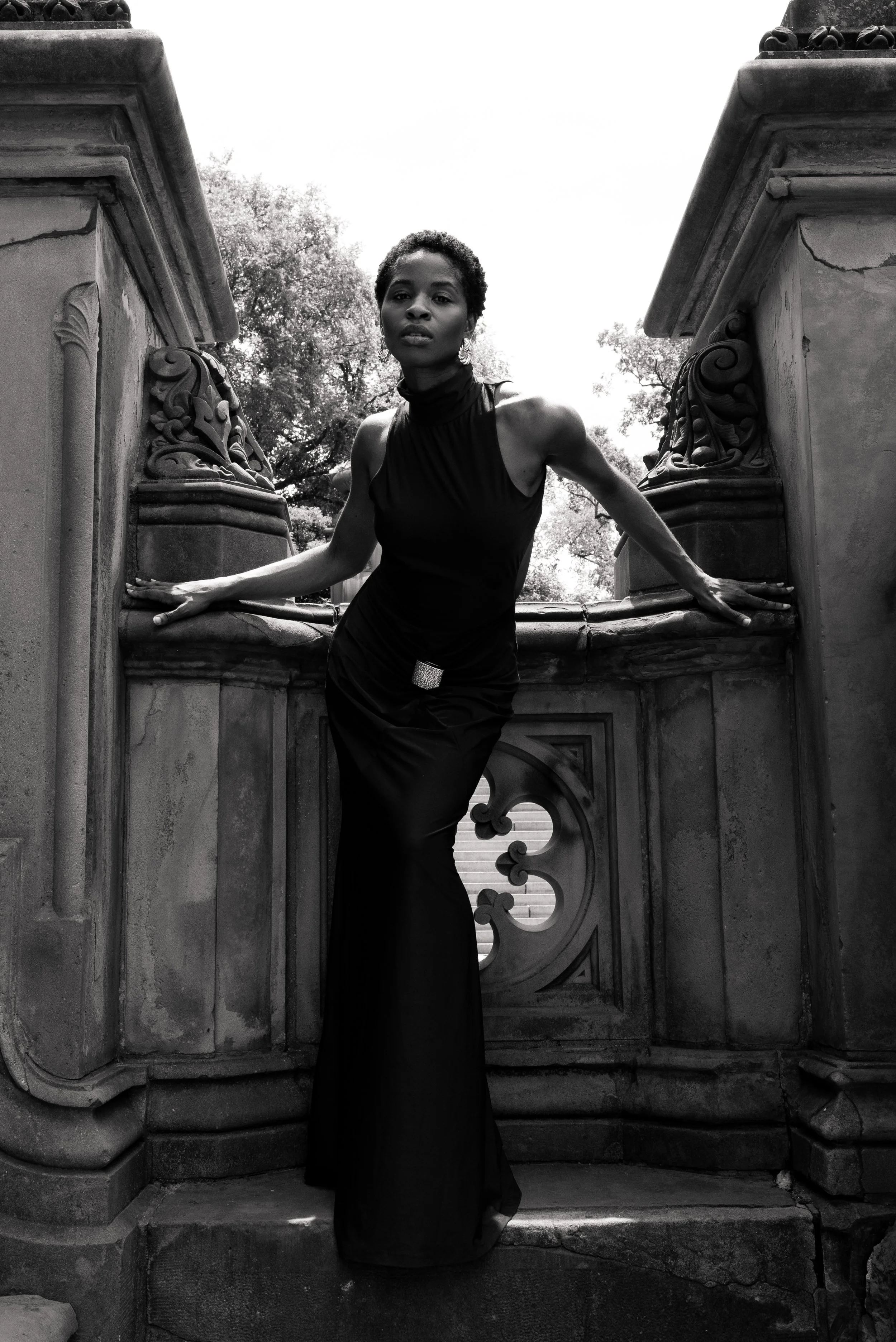 Black and white photo of a woman in a long, elegant dress posing between ornate stone railings on stairs, with trees and sky in the background.