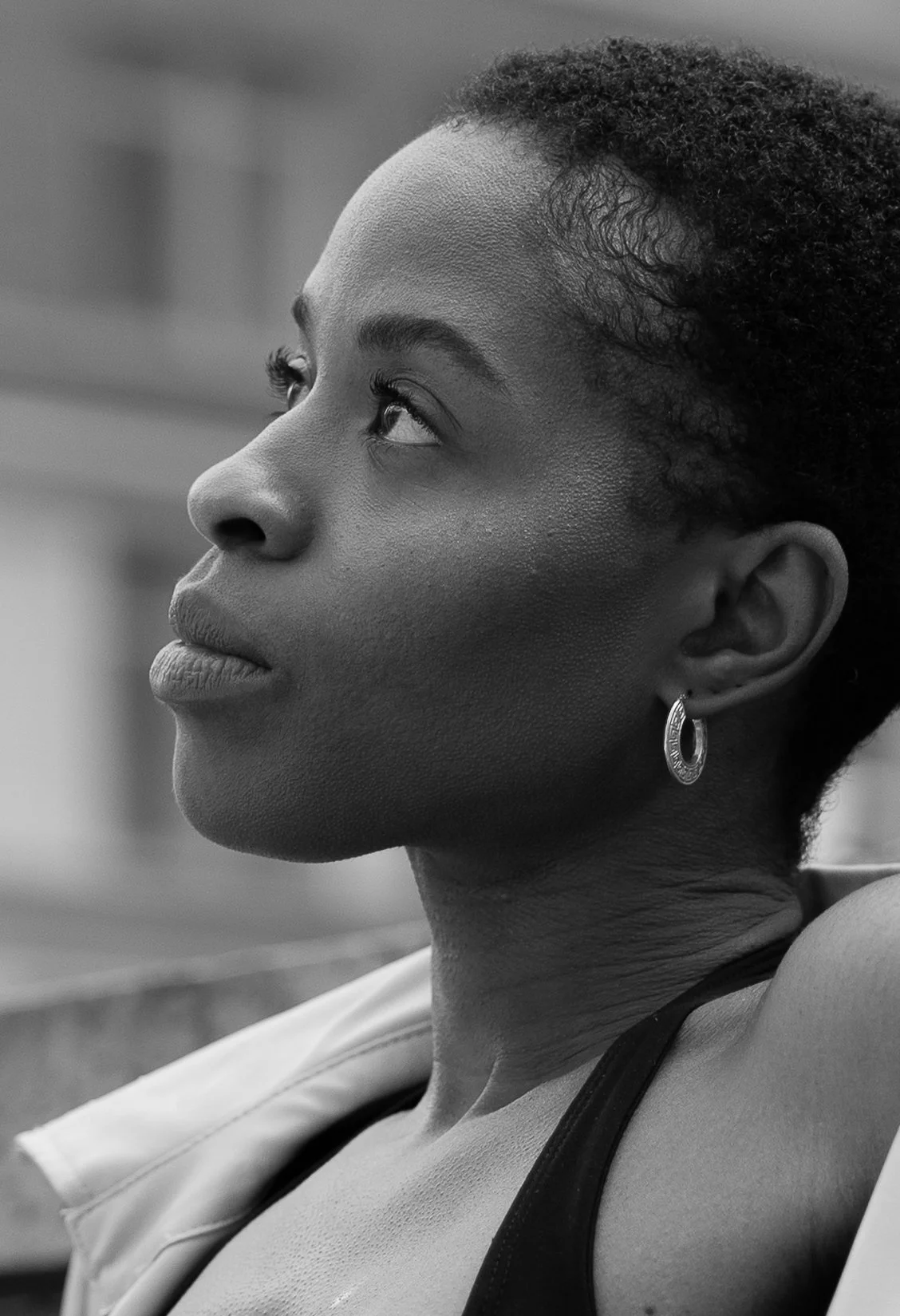 A black and white close-up portrait of a woman with short curly hair, wearing a hoop earring and a tank top, looking upwards.