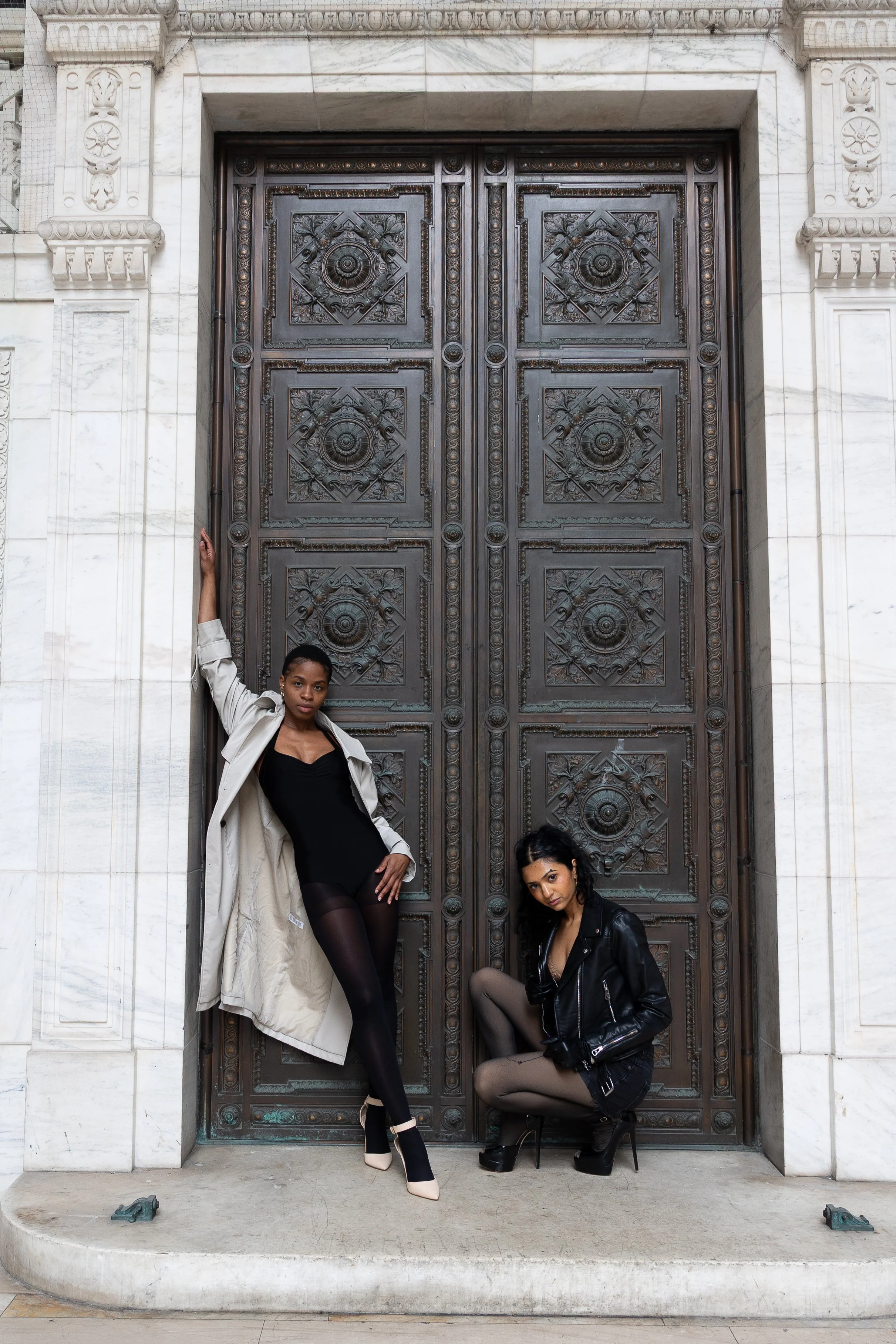 Two women posing in front of large ornate metal door with stone frame, one standing with arm raised and the other squatting.
