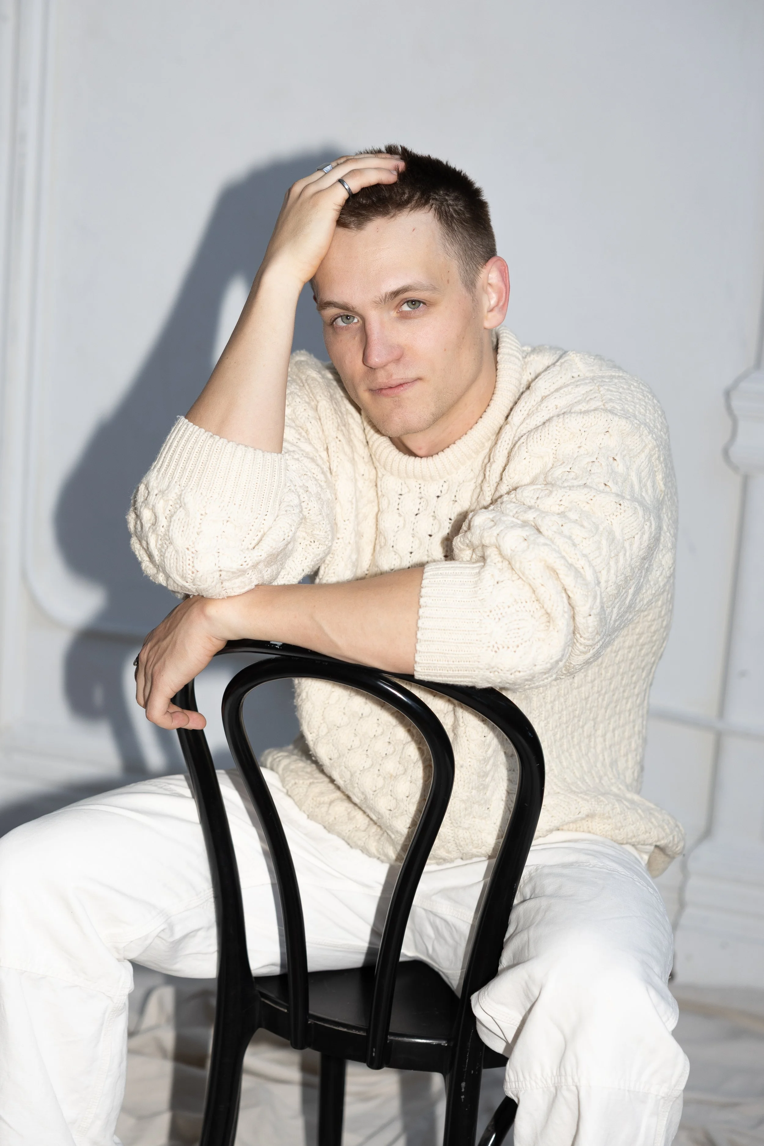 A young man with short dark hair, wearing a cream-colored cable knit sweater and white pants, sitting on a black chair, resting his left arm on the back of the chair, with his right hand touching his head, looking at the camera.