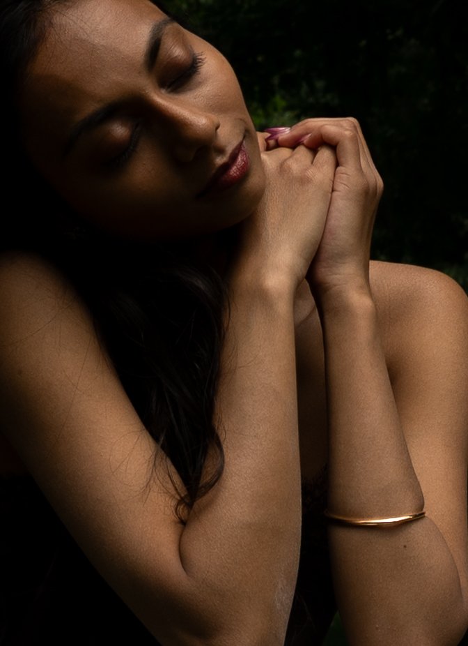 A woman with dark hair and light brown skin, eyes closed, gently holding her chin with her hands, wearing a gold bracelet, and standing against a dark and green background.