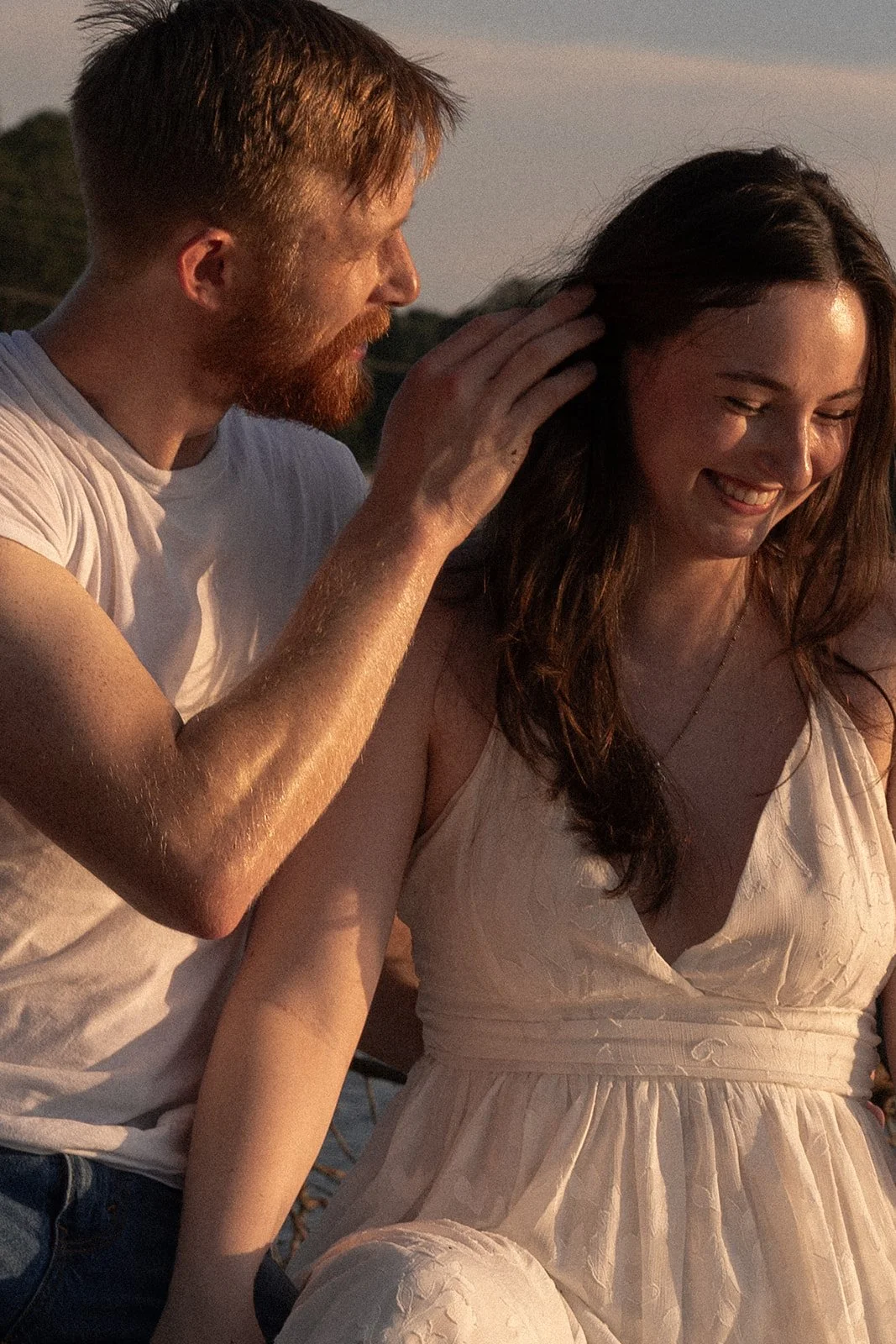 A man is touching a woman's hair during sunset. The woman is smiling and wearing a light-colored dress.