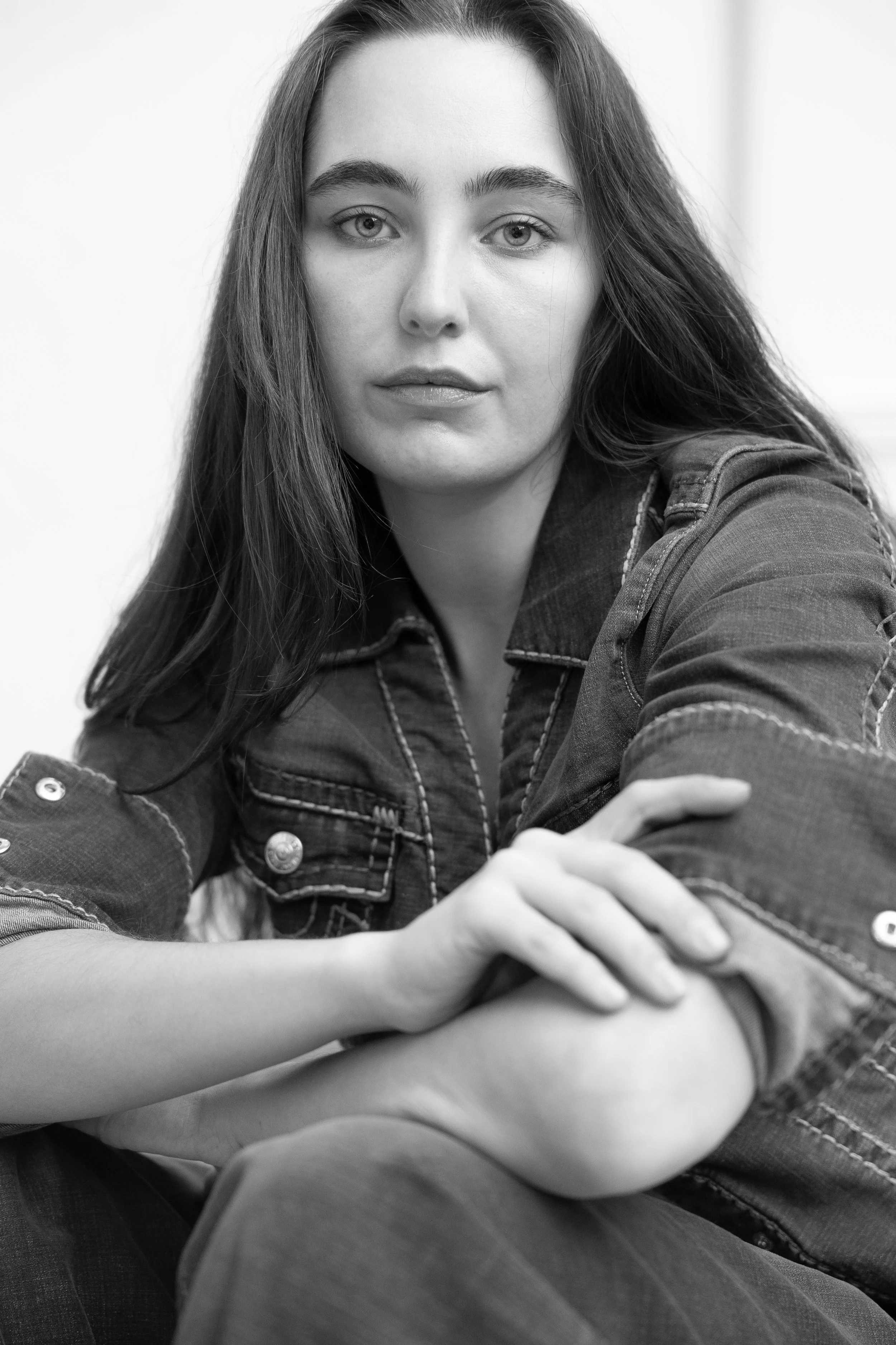 A black and white portrait of a young woman with long hair, wearing a denim jacket, sitting with her arms crossed, looking directly at the camera.
