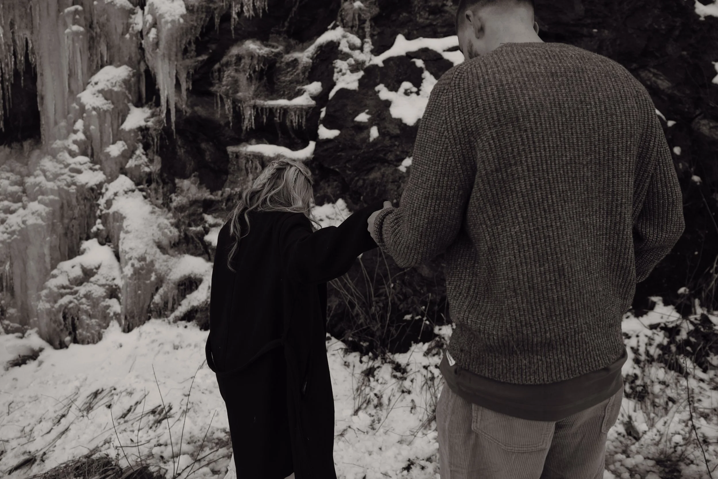 A man and woman in winter clothing holding hands in a snowy outdoor setting near a rocky wall with ice formations.