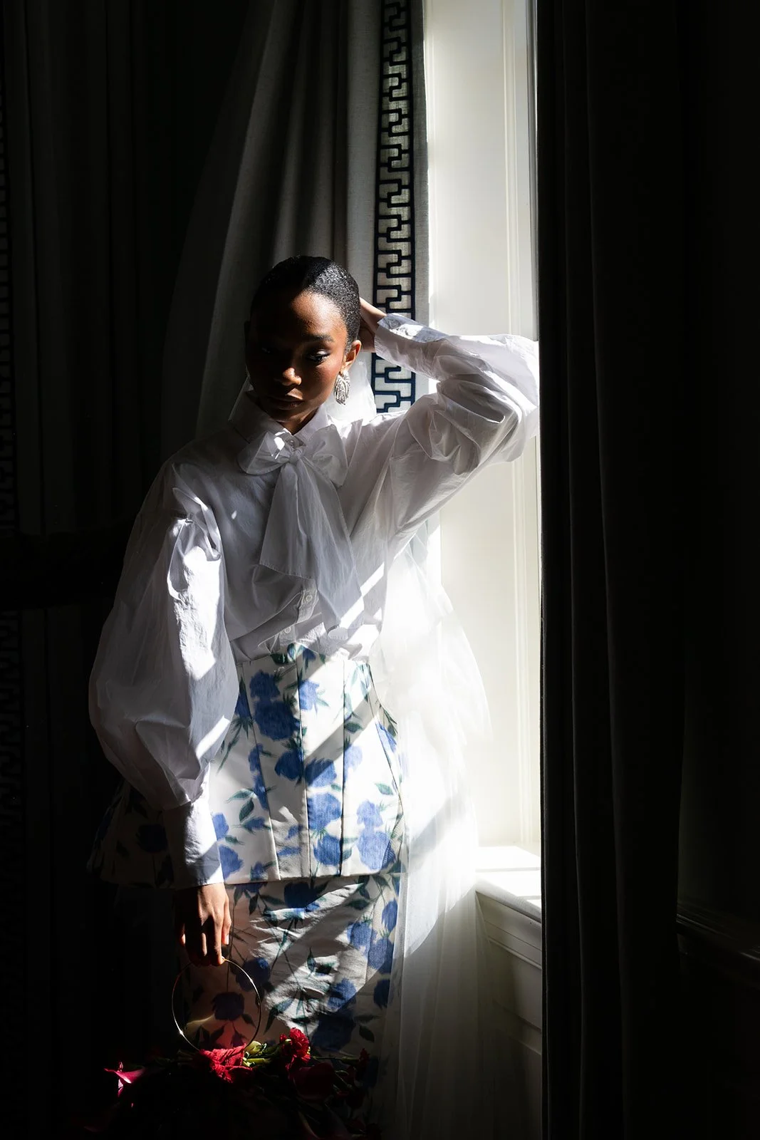 A woman standing by a window with sunlight illuminating her face and white blouse, holding a bouquet of pink flowers.