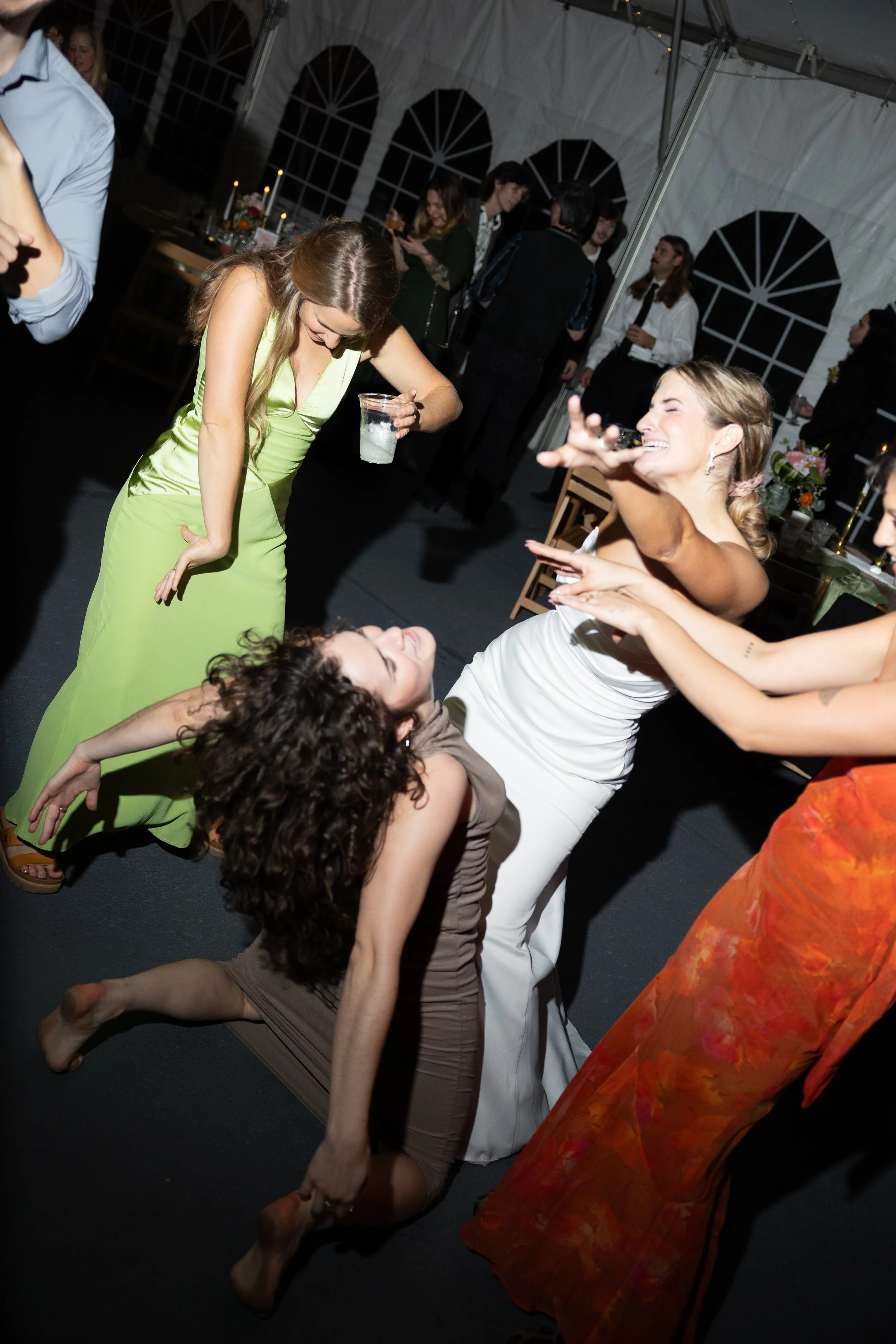 Group of women dancing and having fun at a party in a tented venue, with some women in elegant dresses, smiling and enjoying the moment.