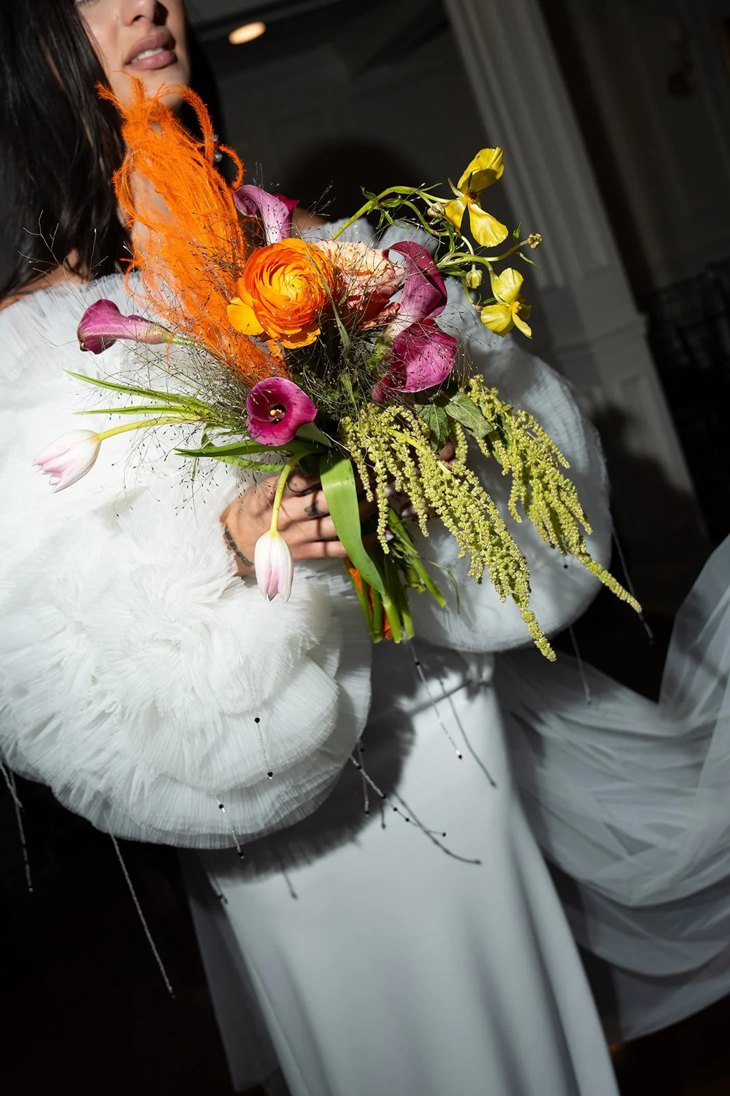 Woman holding a colorful bouquet of flowers, including orange ranunculus, purple calla lilies, pink and white tulips, yellow orchids, and green foliage, inside a decorated room.