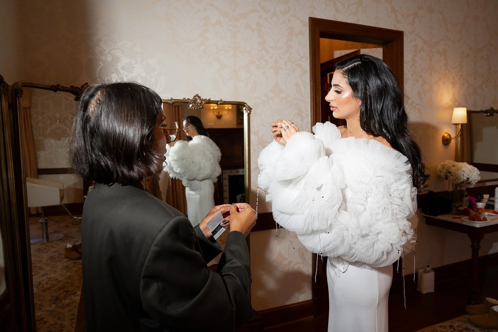 A woman in a white dress with large ruffled sleeves is being assisted by another woman in a black jacket. The woman in white is holding a necklace, and the woman in black is holding a card or label in her hands. They are in a room with a mirror, flor