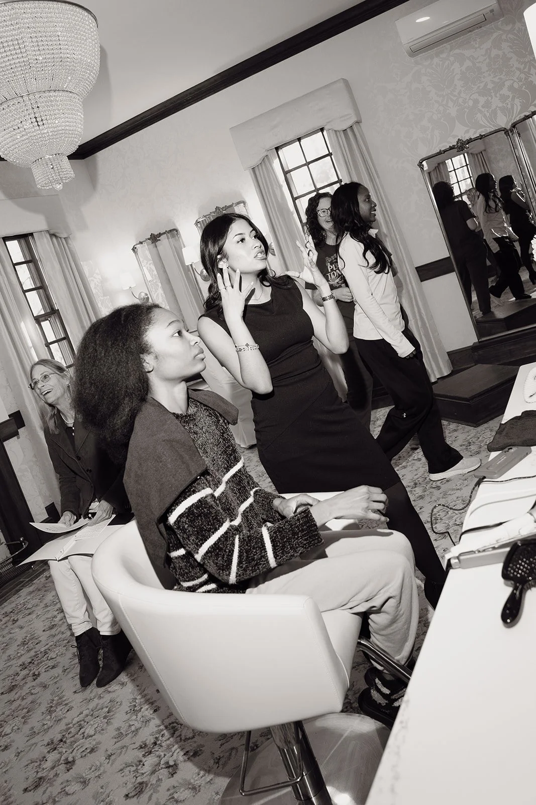 Women in a dressing room, with some standing and others sitting, preparing for a fitting or photoshoot, with large mirrors and a chandelier overhead.