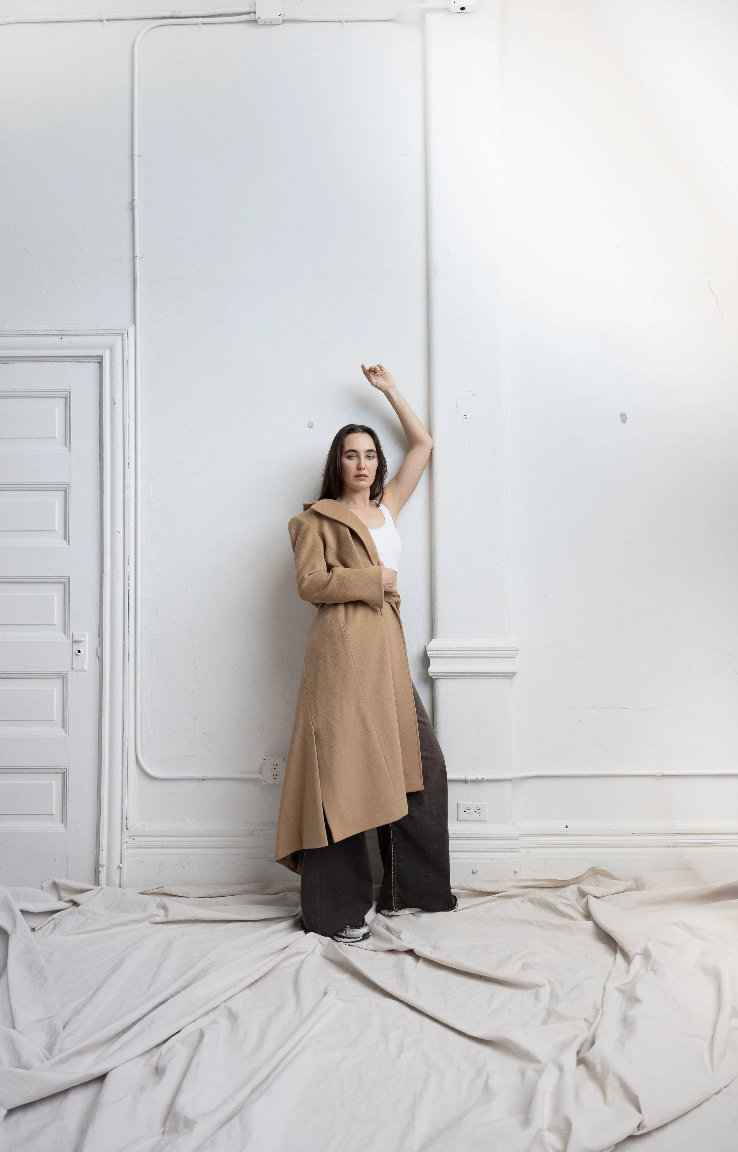 A woman standing against a white wall with her arm raised, wearing a long tan coat, white top, and large dark pants look at the camera.